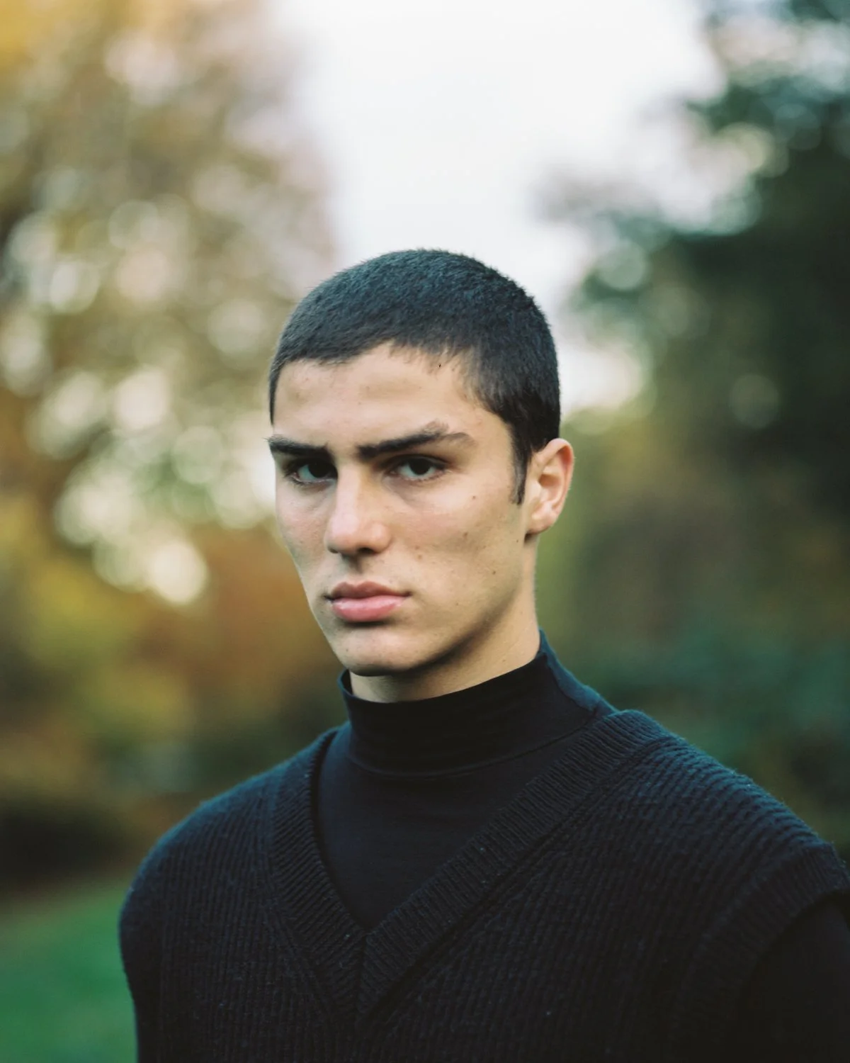 A young man with short dark hair and light skin looking directly at the camera outdoors with blurred autumn trees in the background, wearing a black turtleneck and sweater.