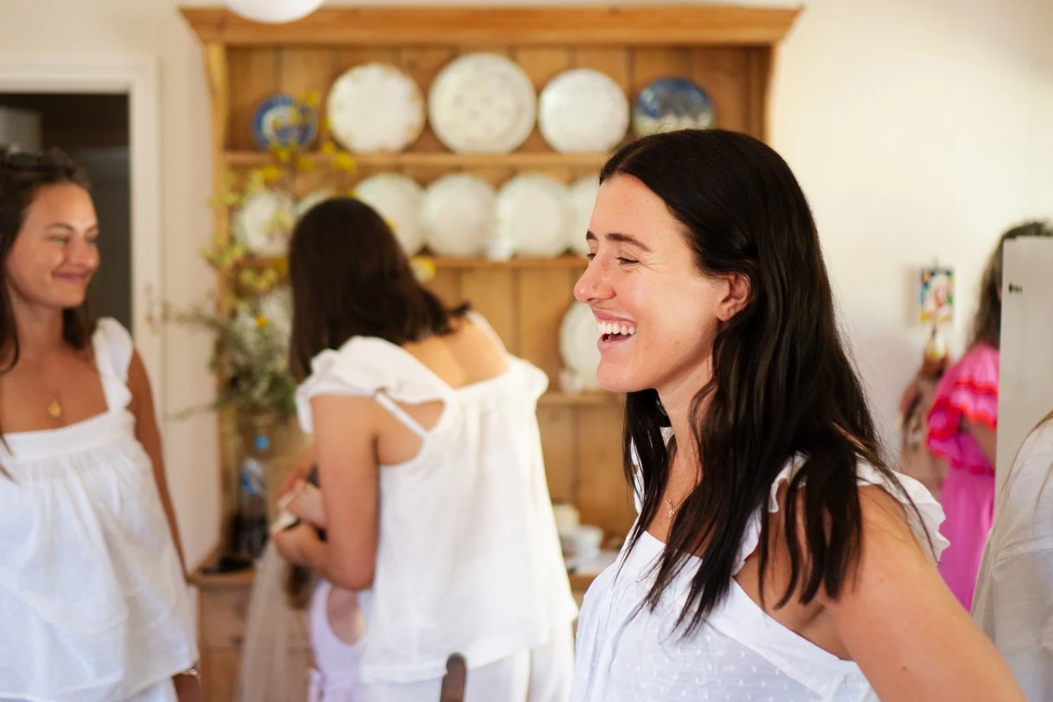 A woman with dark hair smiling in a room with other women, some in white clothes, with a wooden cabinet displaying white plates in the background.