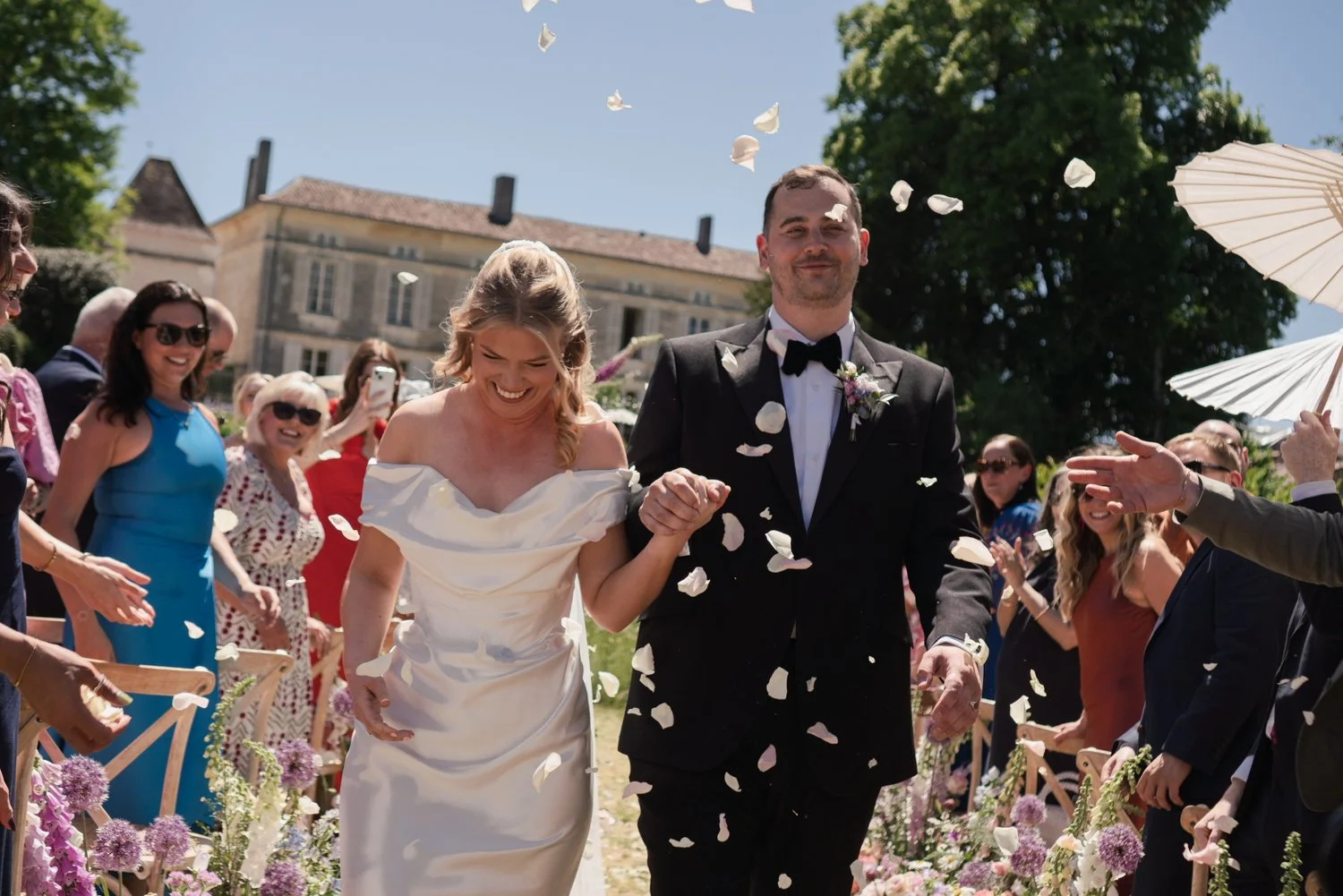 Happy bride and groom walking down the aisle surrounded by guests, with flower petals falling around them at an outdoor wedding on a sunny day.