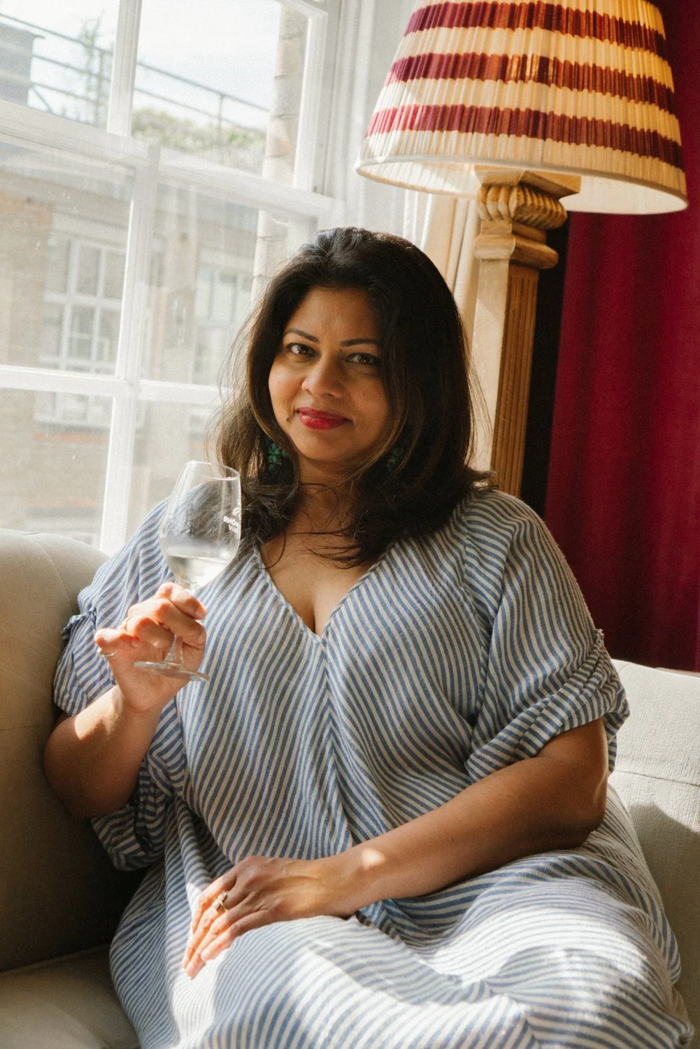 Woman with dark hair and red lipstick sitting on a sofa, holding a glass of white wine, near a window with natural light and a lampshade in the background.
