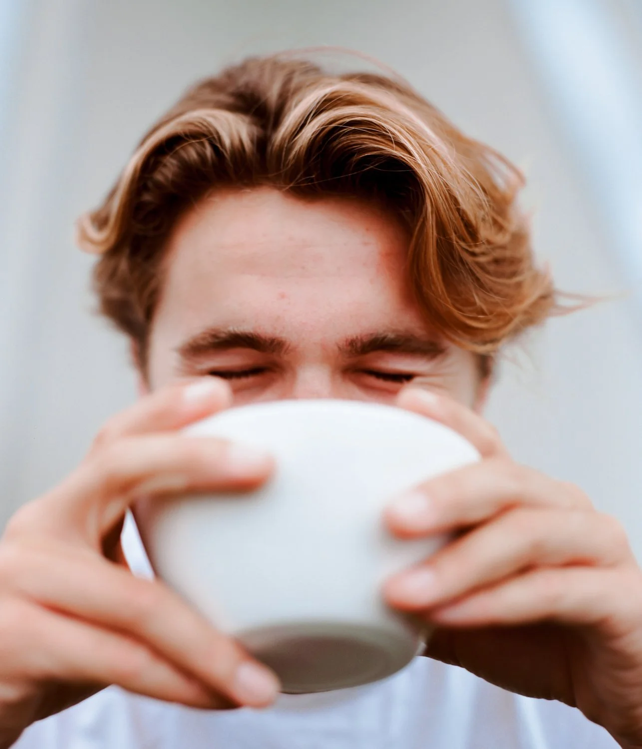 A person with closed eyes and light brown hair drinking from a white cup.