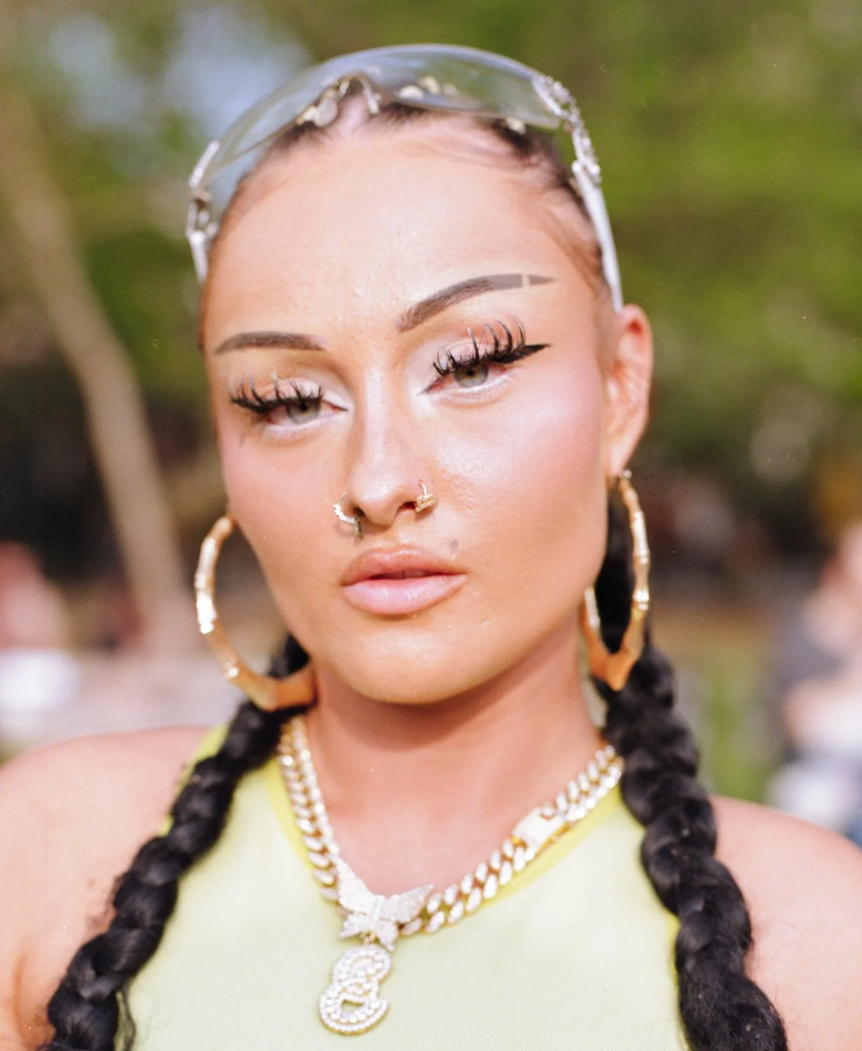 Close-up of a woman with braided hair, wearing large hoop earrings, a nose ring, and a chain necklace, outdoors with blurred trees in the background.