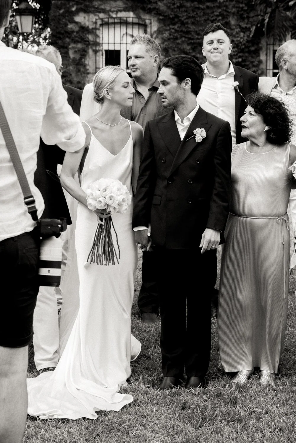 Black and white photo of a wedding scene, with a bride and groom standing close together, surrounded by family and friends outdoors.