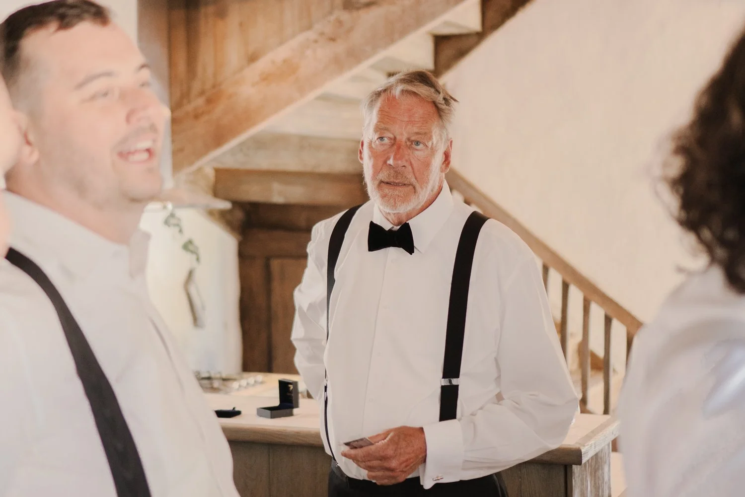 An older man with gray hair and beard, dressed in a white shirt with black suspenders and a bow tie, looking at young men in formal attire inside a rustic wooden building.