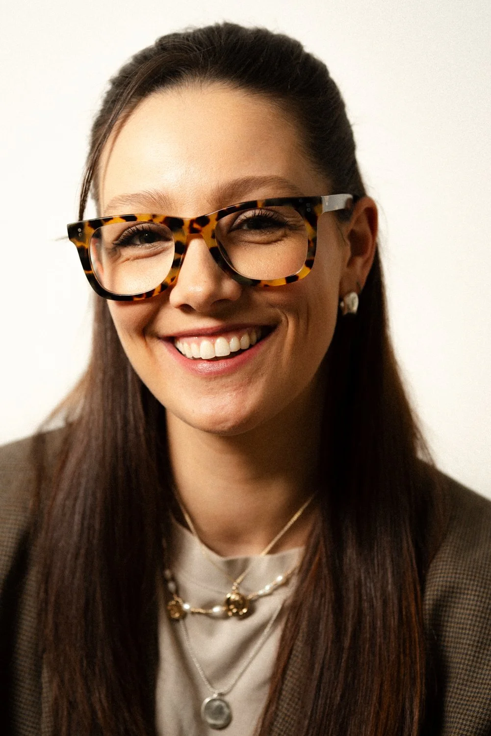 A smiling woman with long brown hair, wearing tortoiseshell glasses, earrings, layered necklaces, a white top, and a brown blazer, against a plain background.