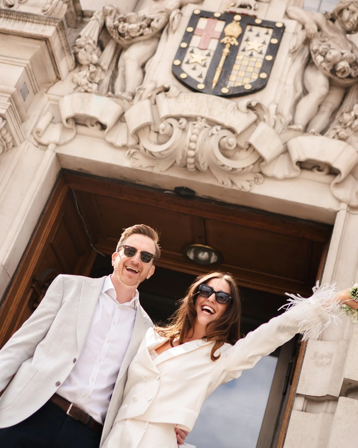 A smiling man and woman in formal attire, wearing sunglasses, standing outside a historic building with a carved stone crest above the entrance.