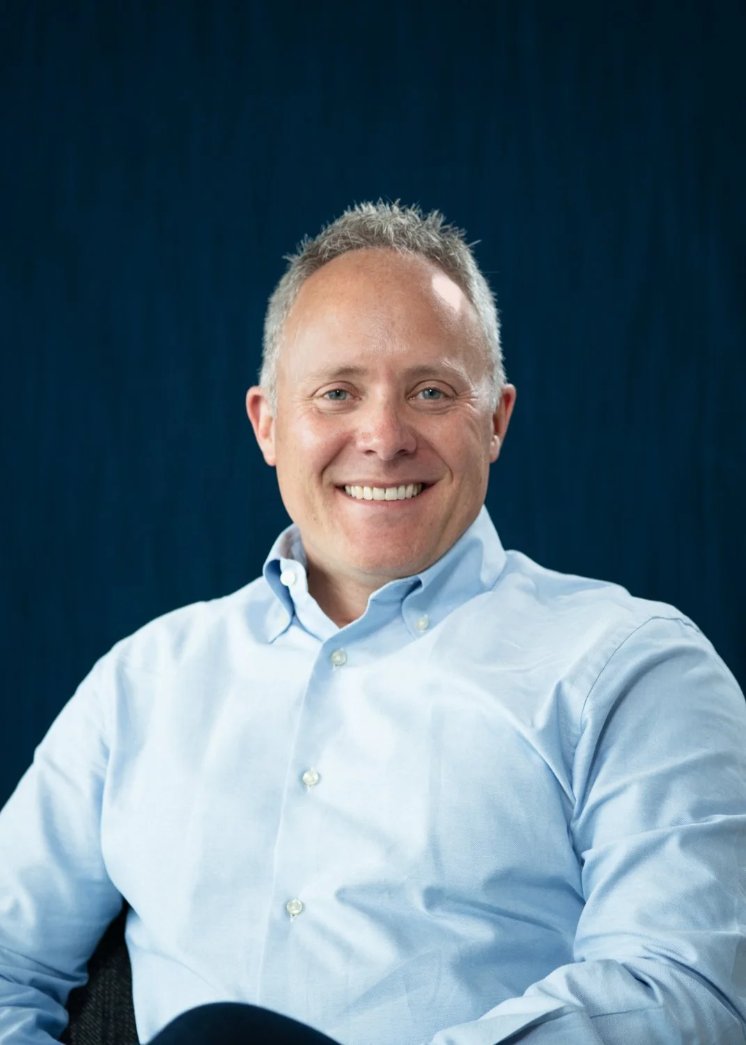 A man with short gray hair, blue eyes, and a big smile, wearing a light blue button-down shirt, sitting against a dark blue background.