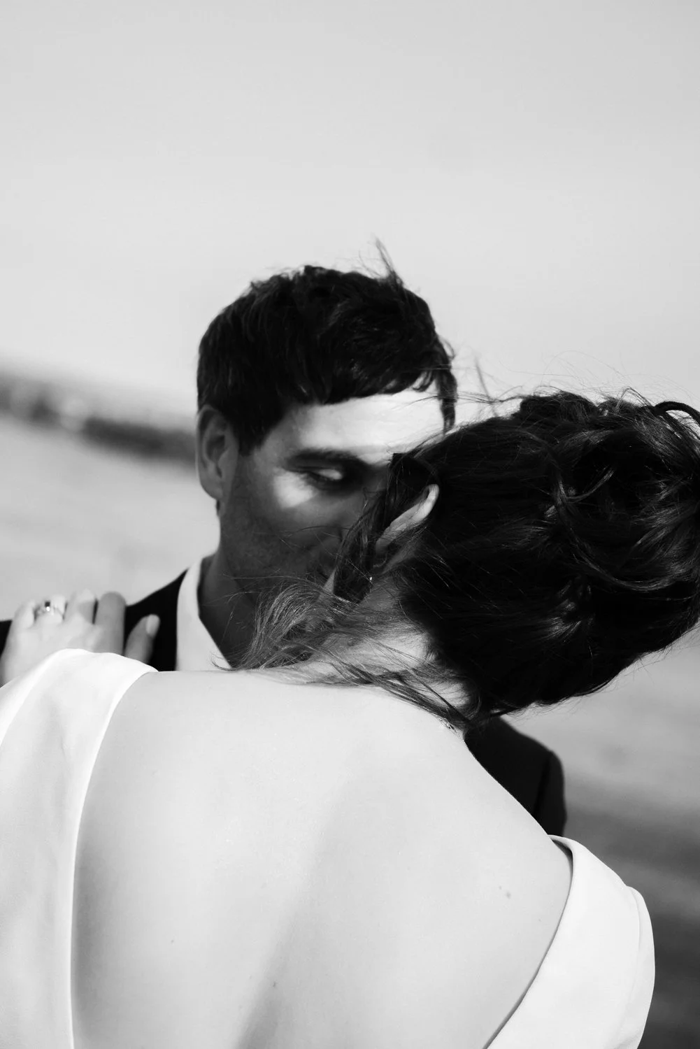 A black-and-white photo of a couple sharing an intimate moment, with their faces close together, outdoors near a body of water.