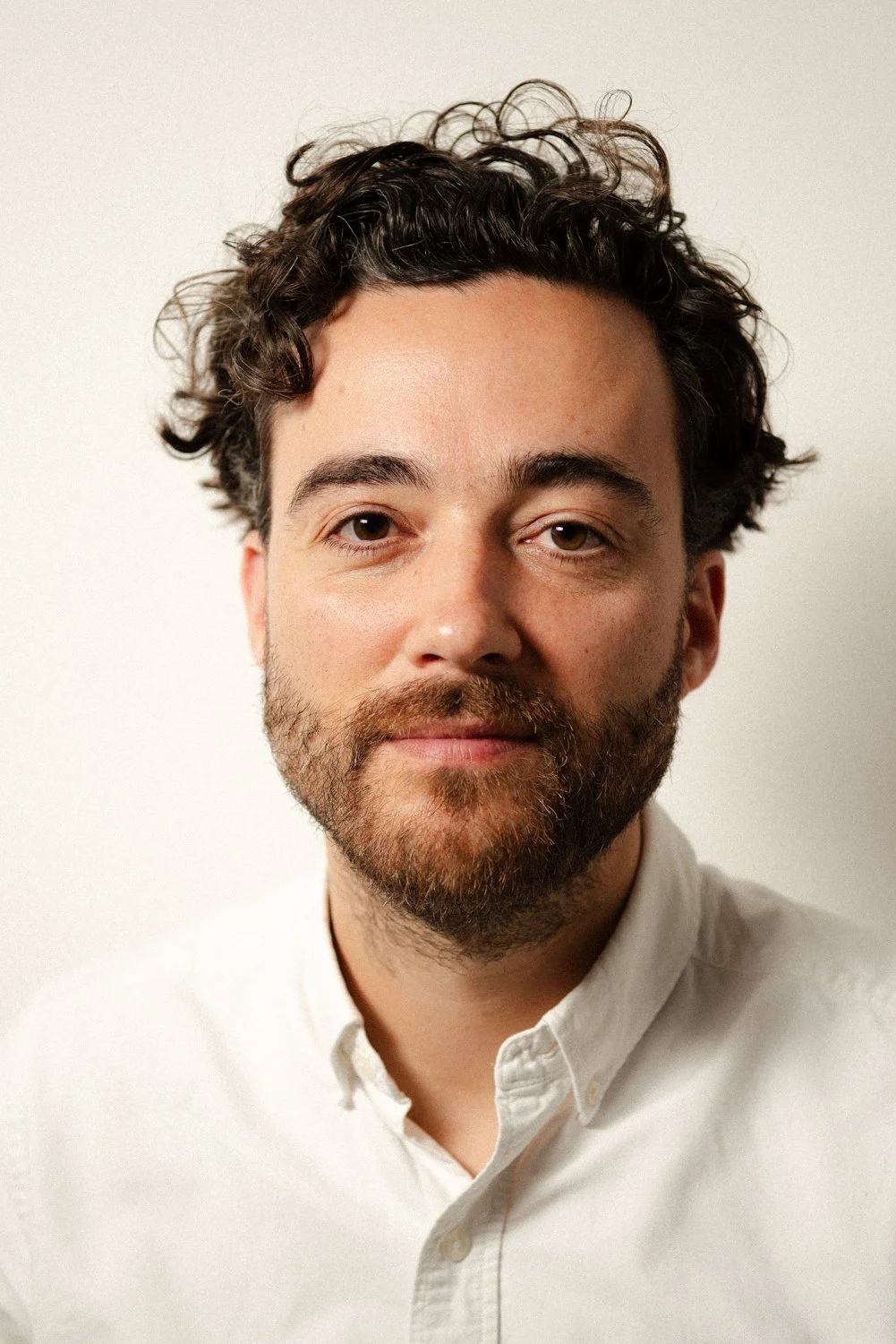 Close-up portrait of a man with dark curly hair, a beard, wearing a white collared shirt, against a plain light background.