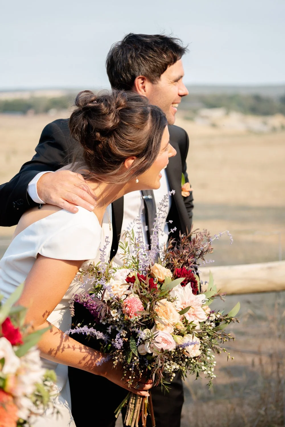 Happy couple celebrates wedding outdoors, holding a bouquet of flowers, in a scenic rural landscape.