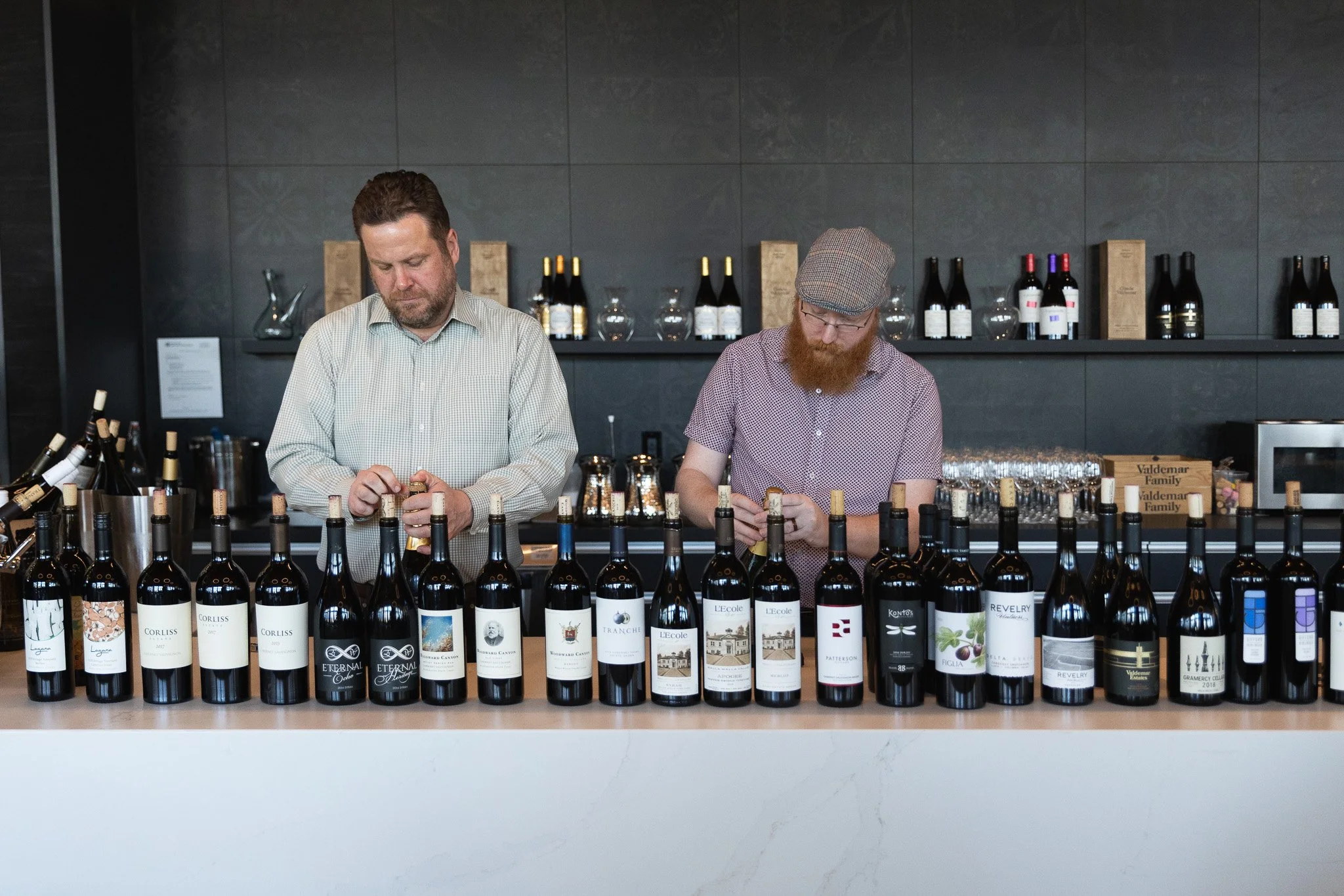 Two men are standing behind a counter with multiple bottles of wine, inspecting or preparing them, in a modern wine tasting room or store.