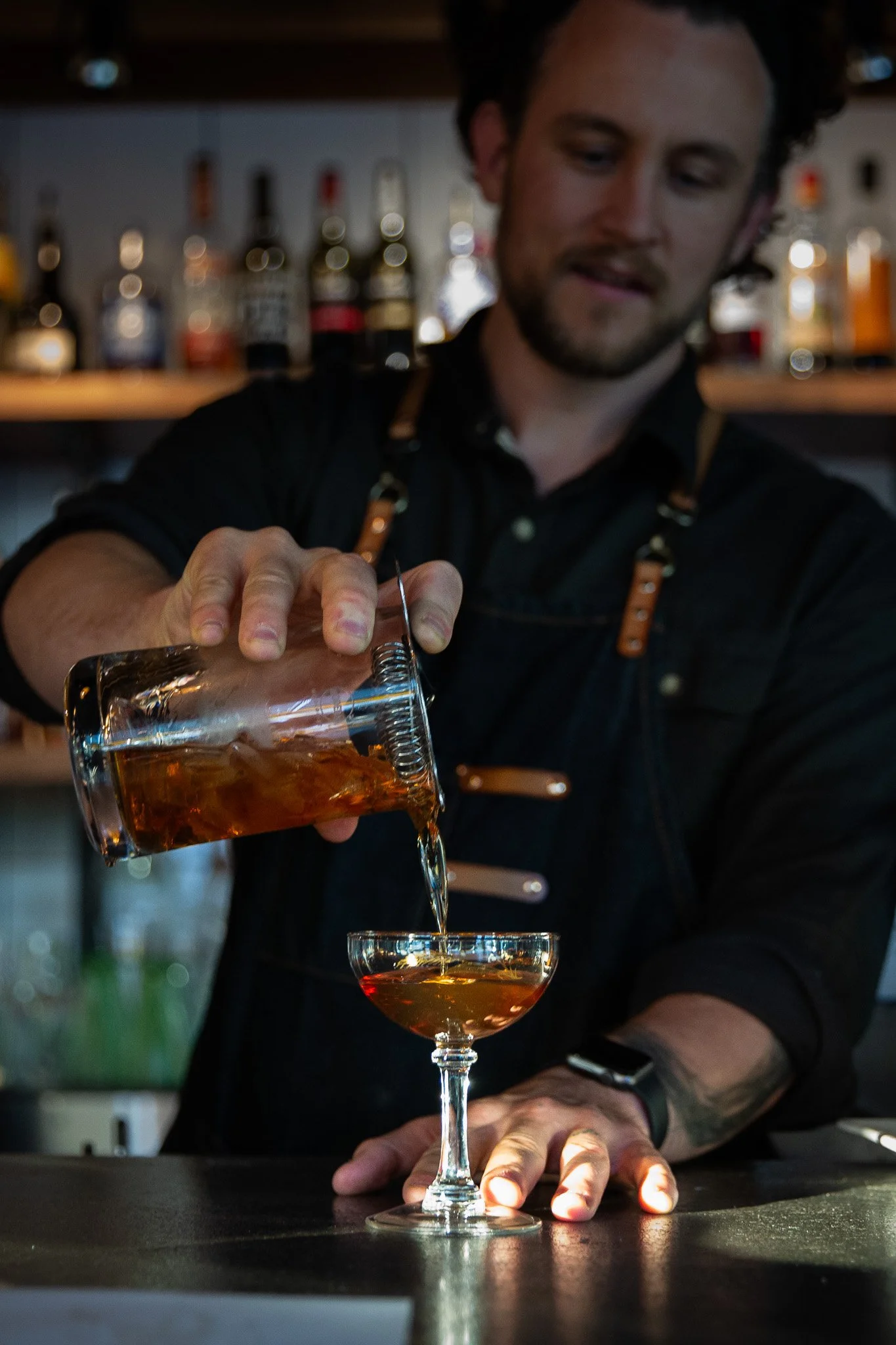 Bartender pouring a drink from a glass container into a cocktail glass at a bar.