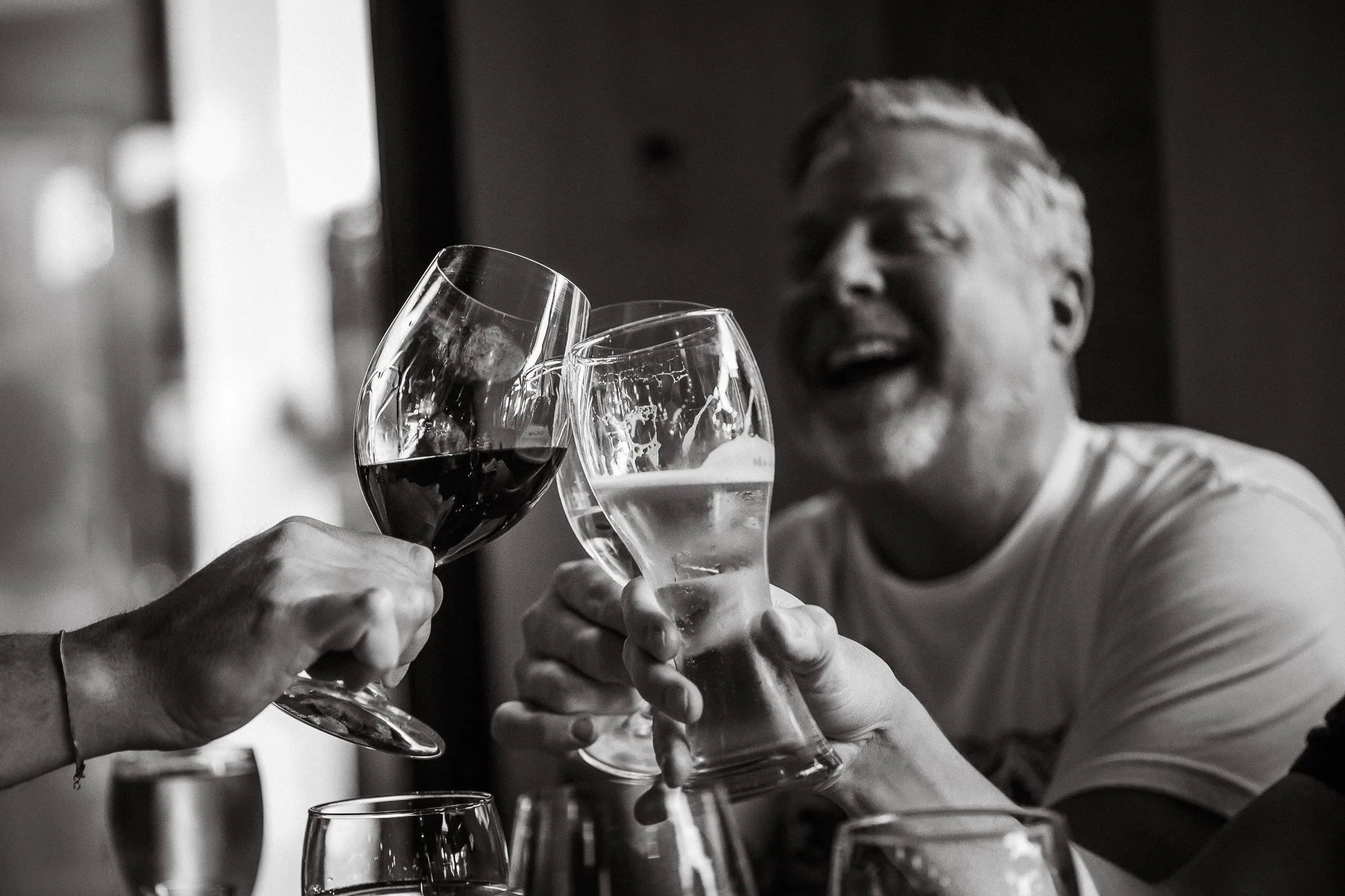 Two people clinking glasses, one with red wine and the other with a clear beverage, in a social setting. The background shows a smiling man with a beard and short hair.