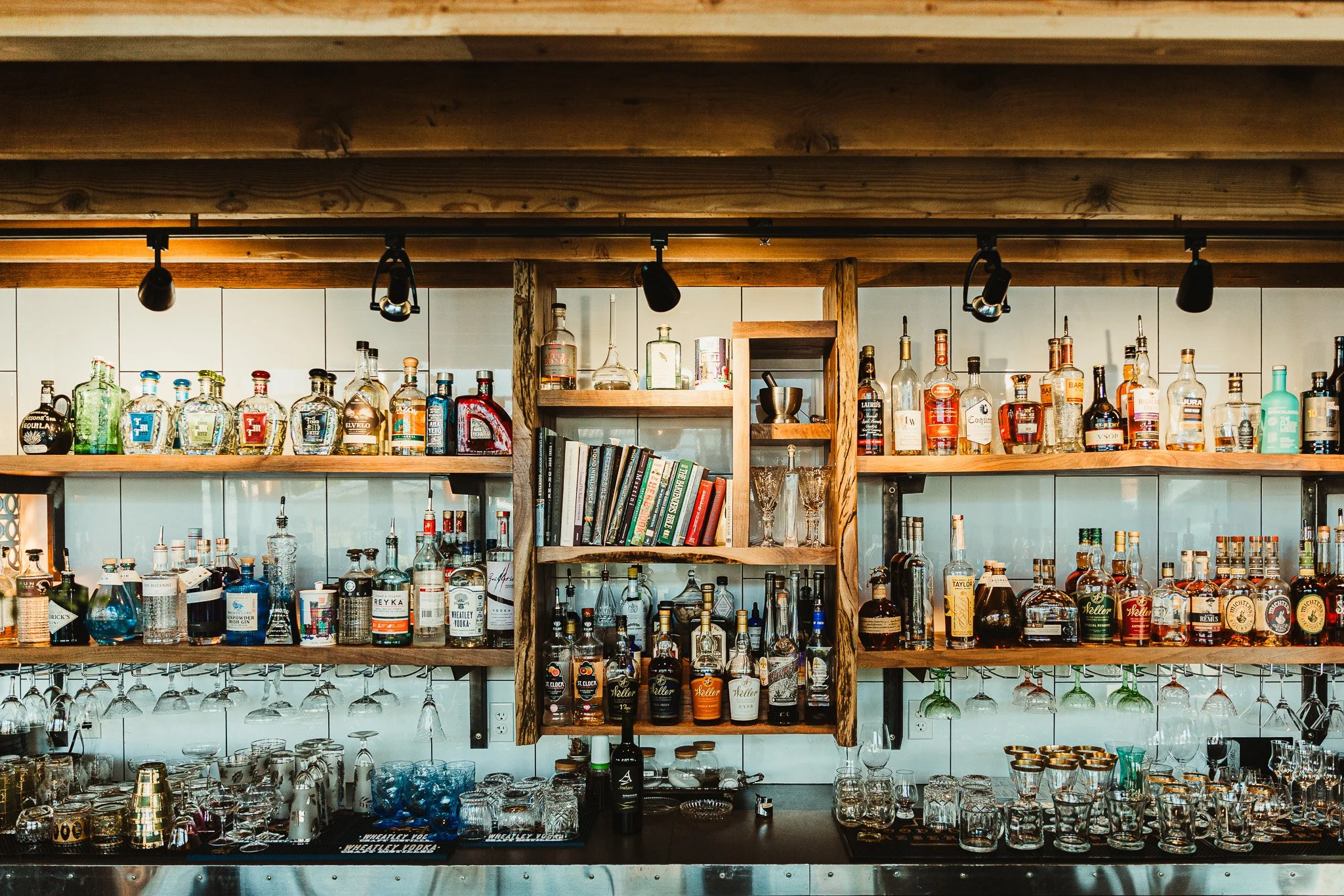 Bar with shelves of various liquor bottles, glasses, and books, illuminated by ceiling lights.