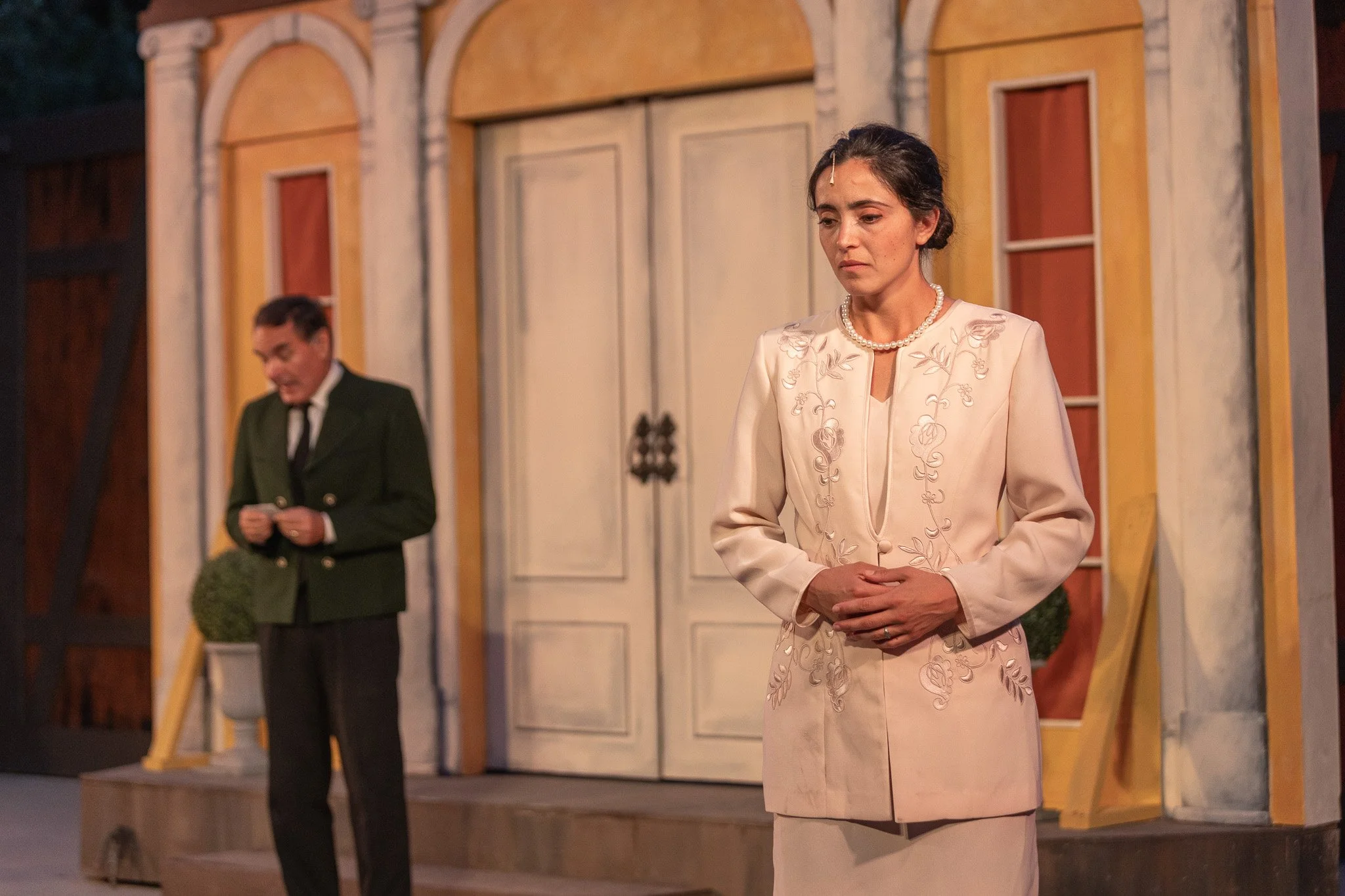 A woman with dark hair in an elegant white suit with embroidery, wearing a pearl necklace, stands with hands clasped in front of her, looking downcast. Behind her, a man with dark hair in a green jacket and black pants is reading a note or paper, set