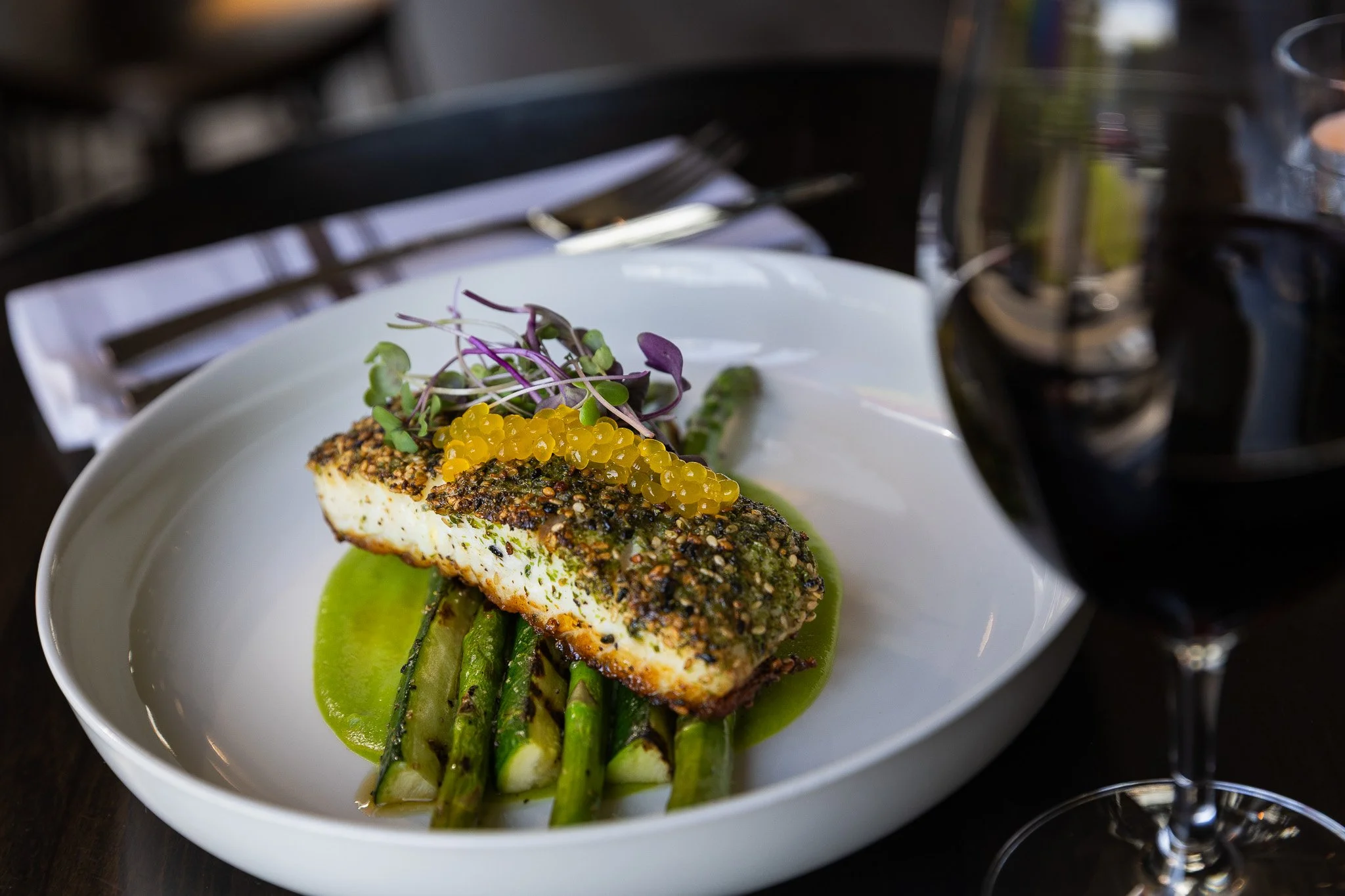 A plated seafood dish featuring a crusted fish fillet, grilled asparagus, a green sauce, microgreens, and yellow caviar, with a glass of red wine nearby.