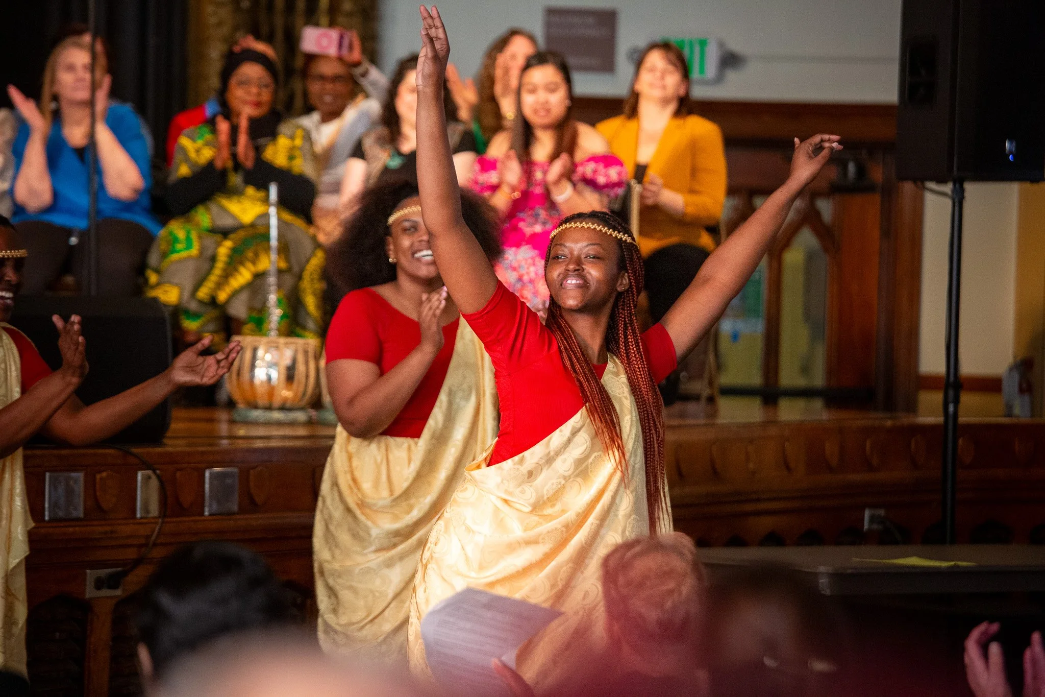 A woman in a traditional outfit with a gold sash and red top dancing with arms raised, surrounded by applause and an audience of diverse women, in an indoor event setting.