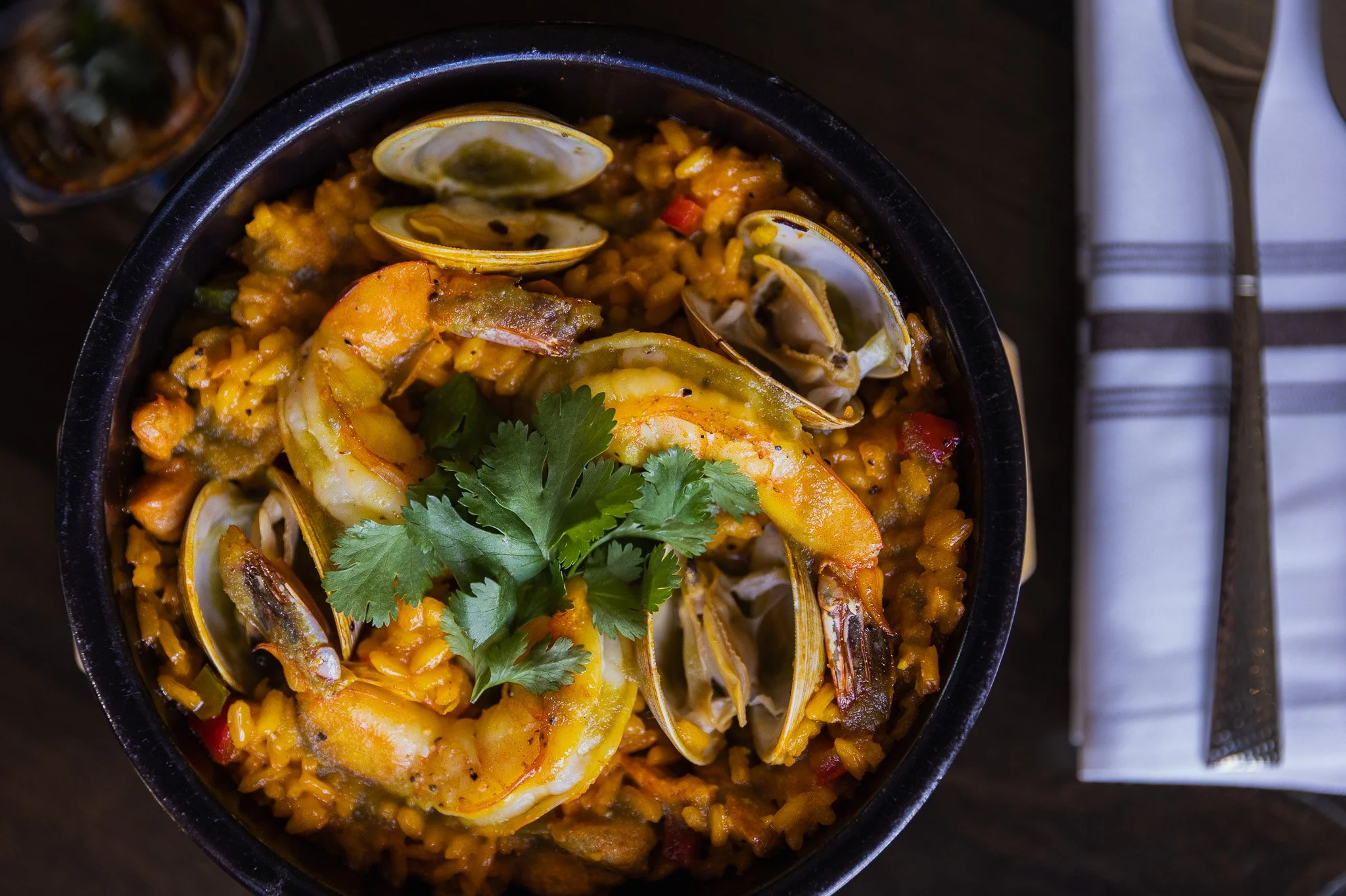 Seafood paella with shrimp, mussels, rice, and garnished with cilantro in a black bowl on a dark wooden table.