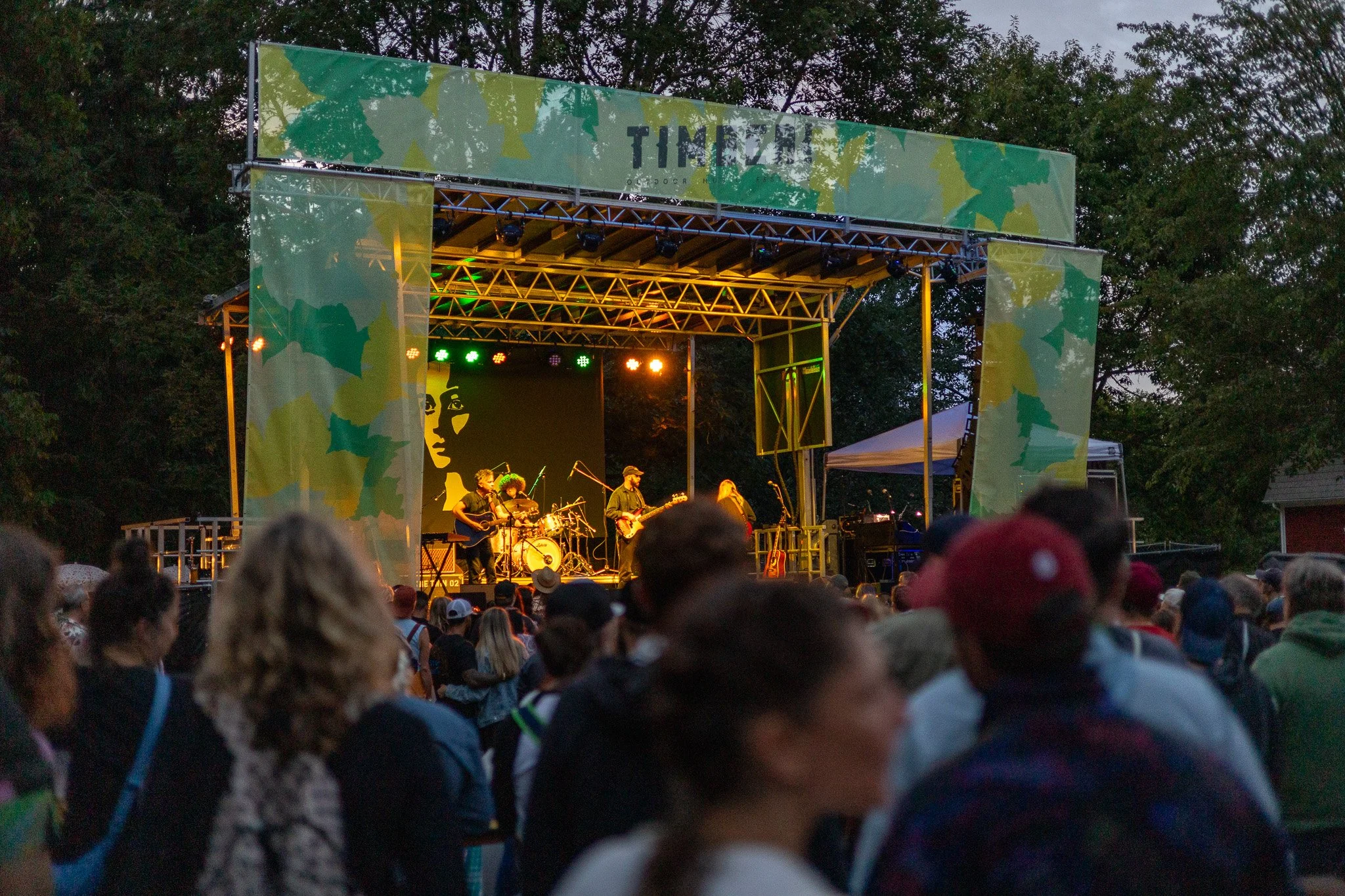 Outdoor concert with a band performing on stage, audience gathered in front, trees in the background, evening setting.