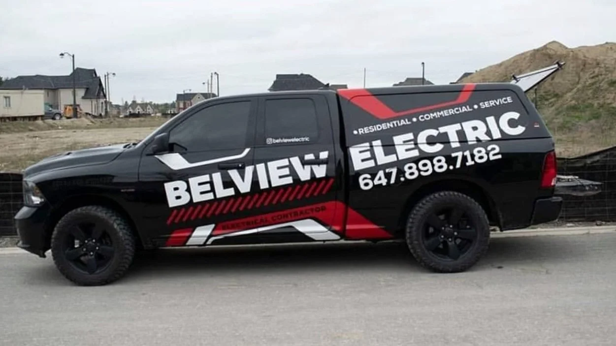 A black pickup truck with the company name 'Belview Electric' and contact info in white and red lettering, parked on a construction site with houses and dirt piles in the background.
