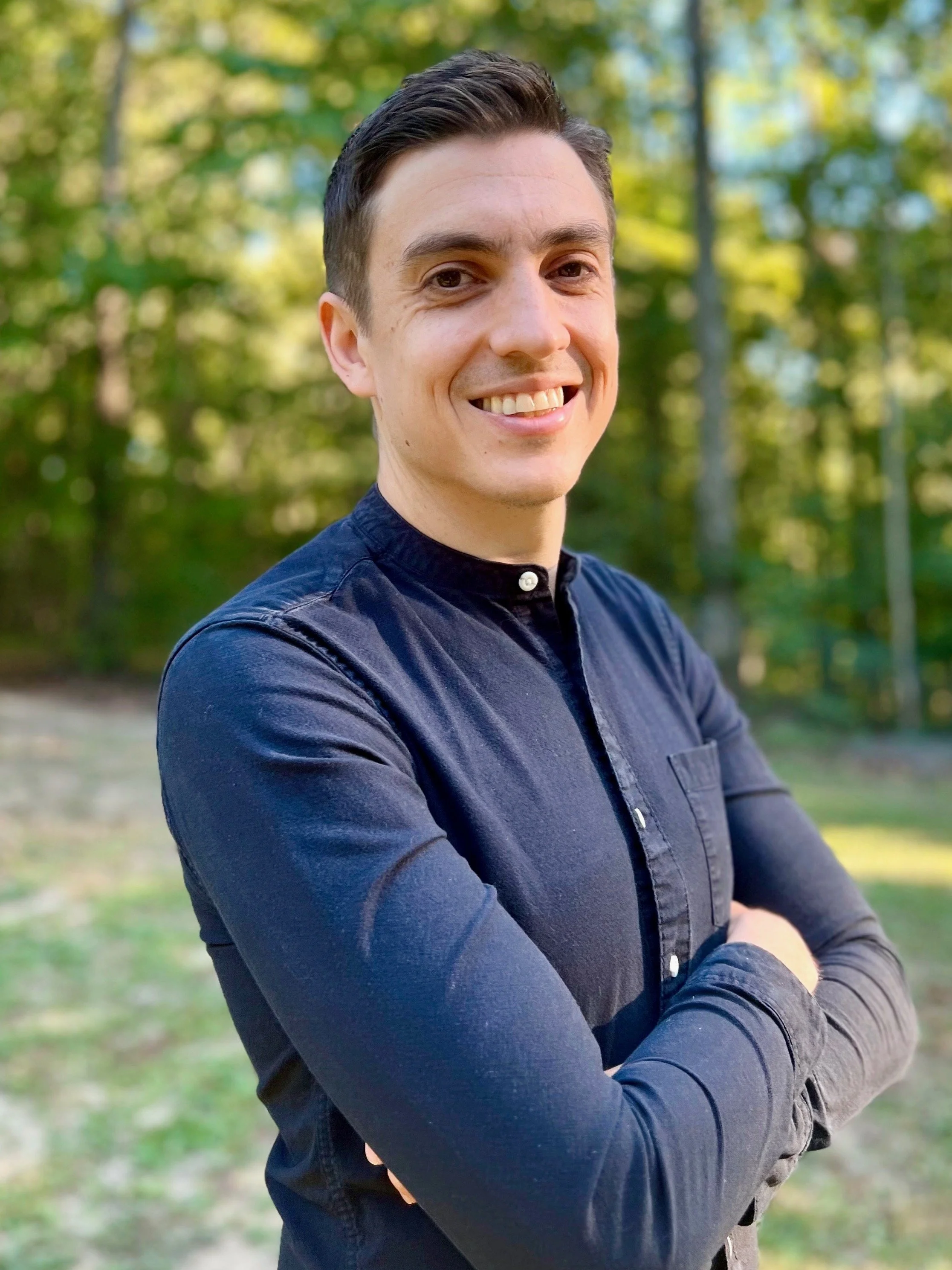 A young man smiling outdoors with arms crossed, wearing a dark blue shirt, standing in a green wooded area.