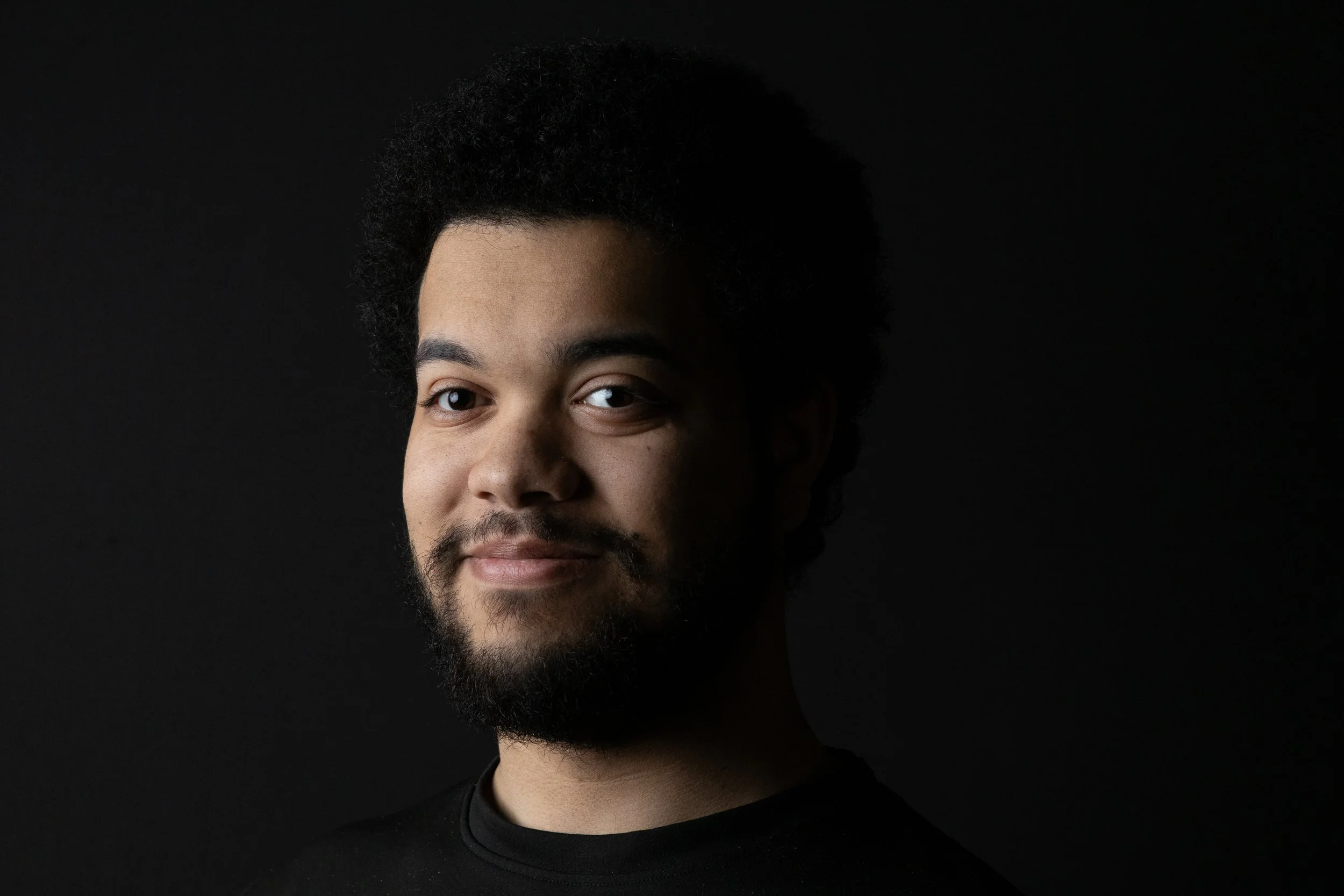 A portrait of a young man with curly hair and a beard, smiling slightly, against a dark background.