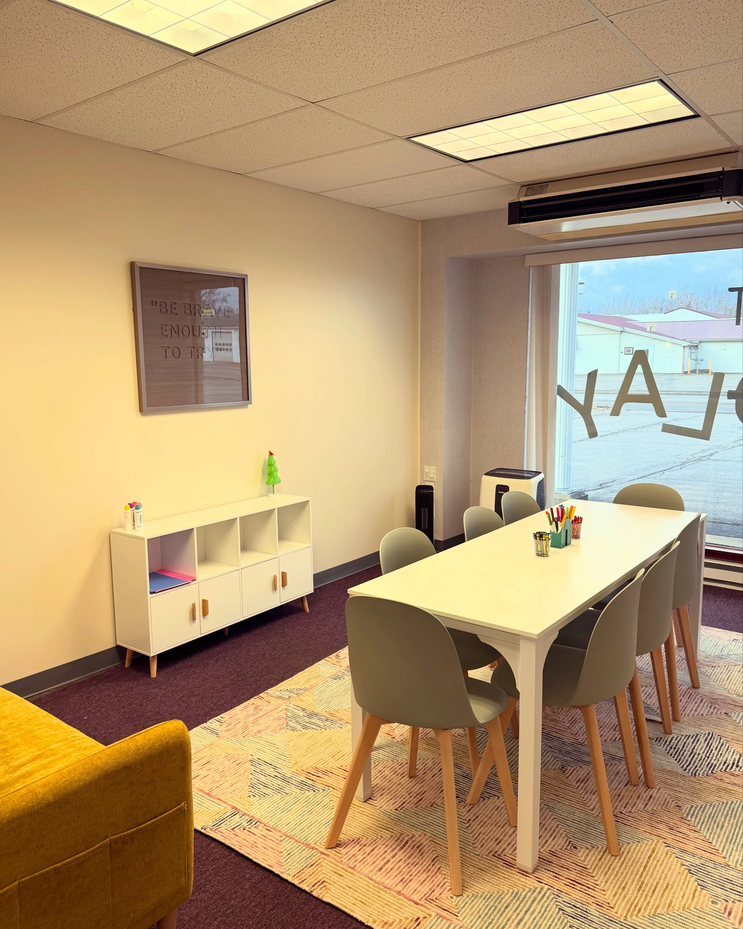 A brightly lit classroom with a white rectangular table and six light gray chairs around it. On the table are cups filled with colored markers. To the left, a white cabinet with three open cubbies and four closed drawers beneath. A framed quote on the yellow wall reads "BE BRAVE ENOUGH TO TRY." The large window on the right shows an outdoor parking lot and other buildings. The room has a colorful geometric rug, purple carpet, and a yellow armchair partially visible in the foreground.