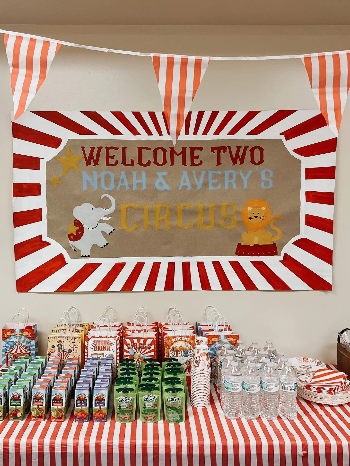 Circus-themed party table with gifts, juice boxes, water bottles, and red and white striped decorations, featuring a sign that says "Welcome Two Noah & Avery's Circus.".