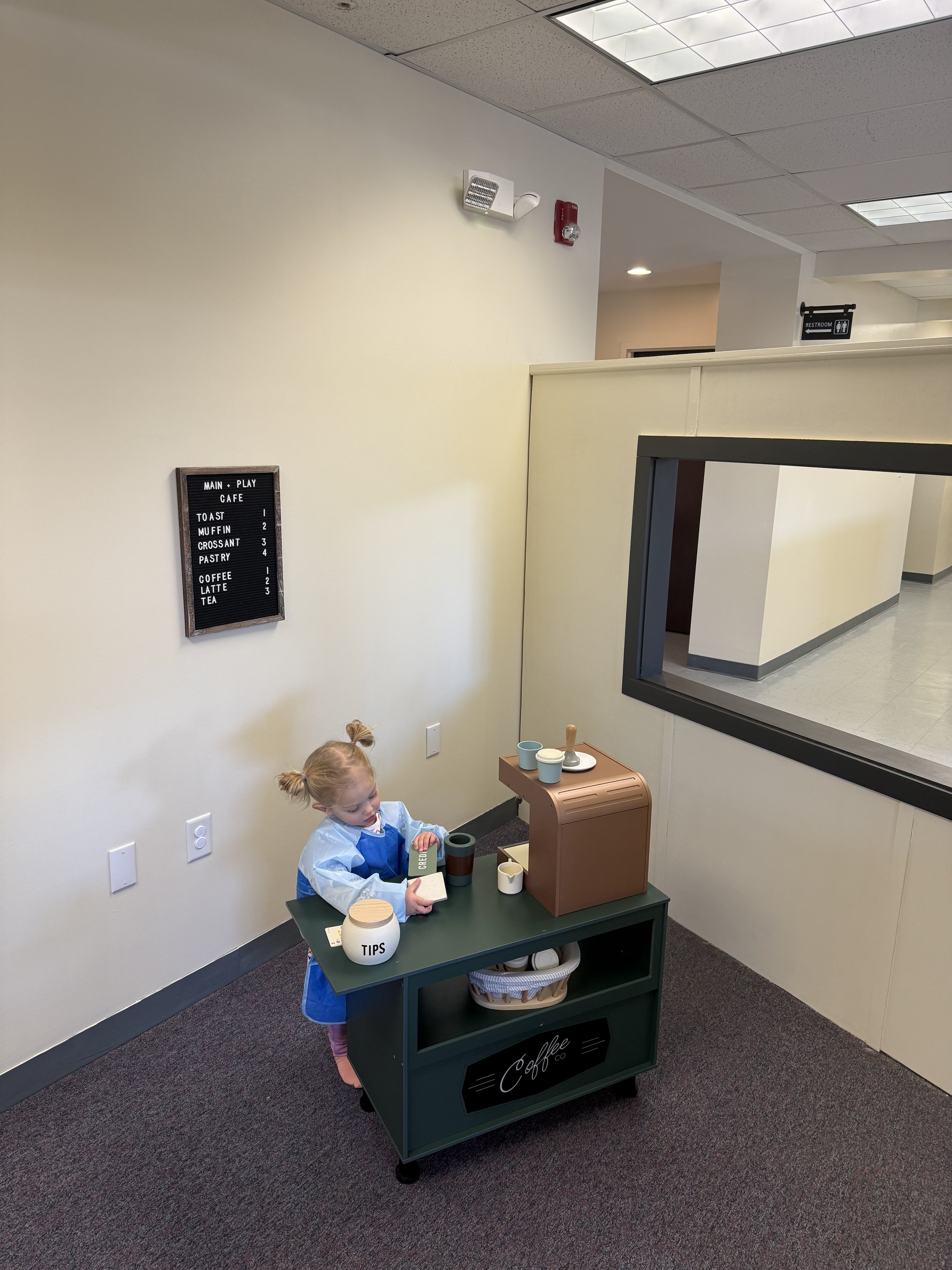 Child playing cafe in a toy kitchen set with pretend coffee machine, cups, and tips jar, in an indoor play area.