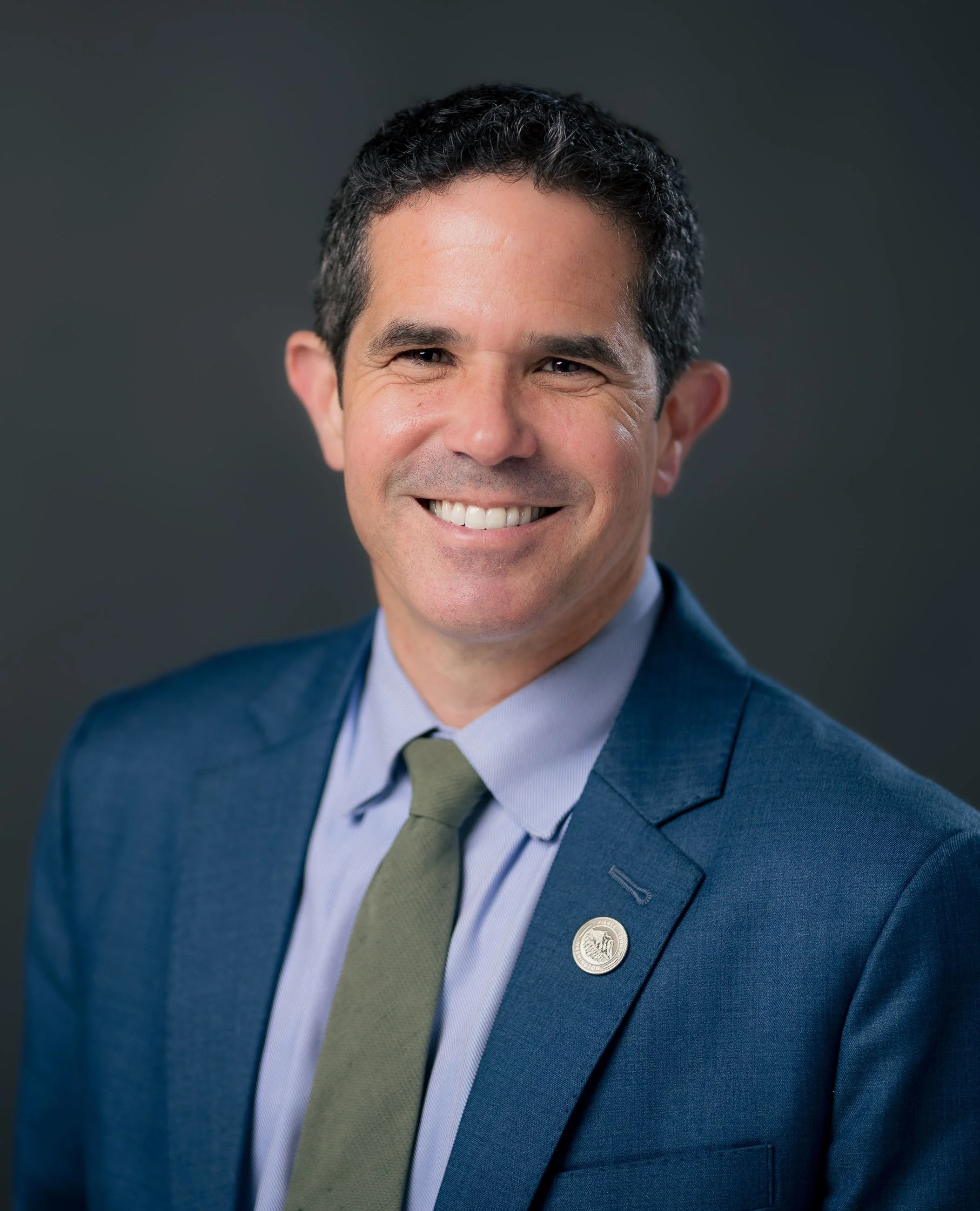 Professional headshot of a smiling man wearing a blue suit, light blue dress shirt, and green tie, against a dark grey background.
