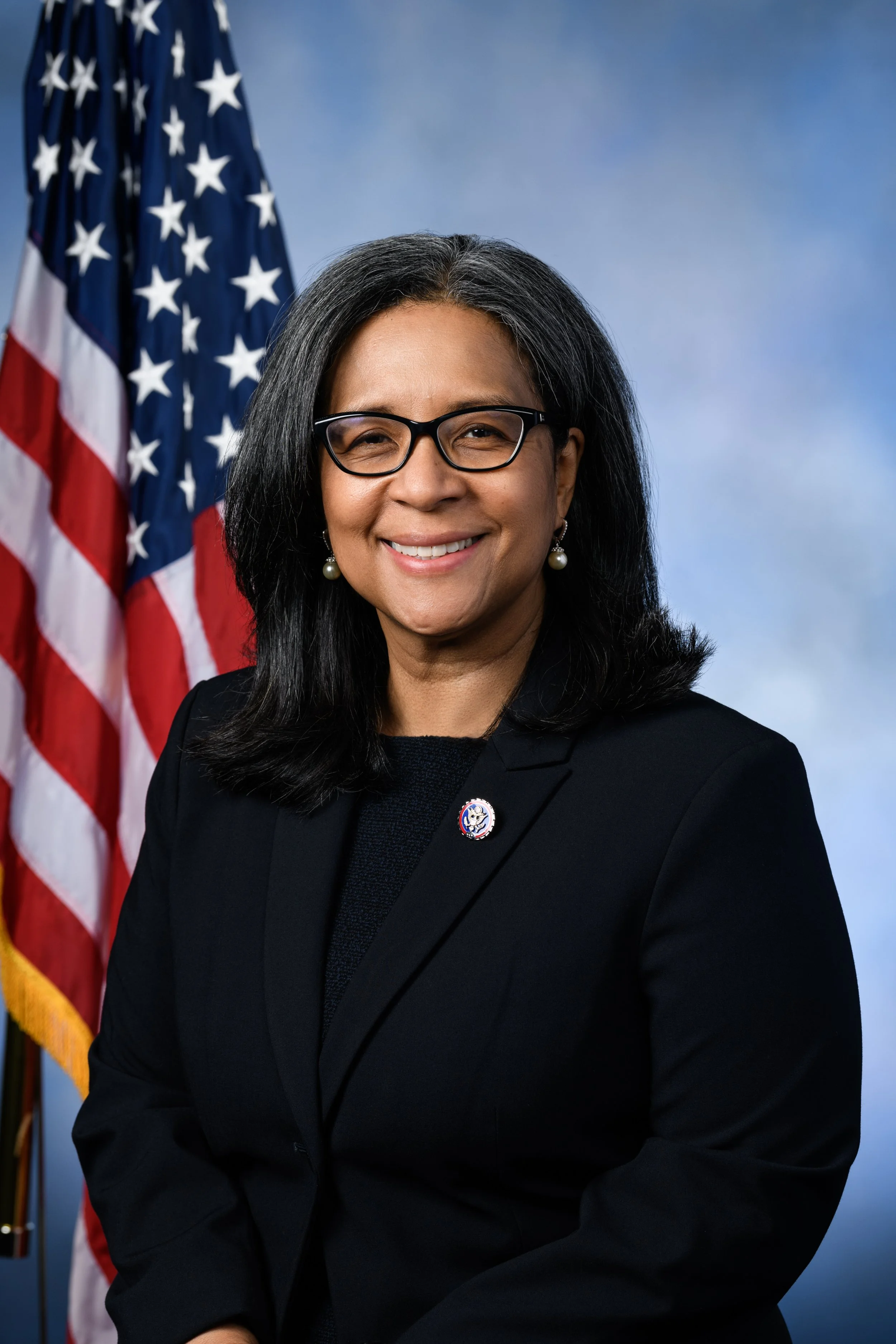 A woman with black hair, glasses, and pearl earrings, wearing a black blazer, standing in front of American flags, smiling for a professional portrait.