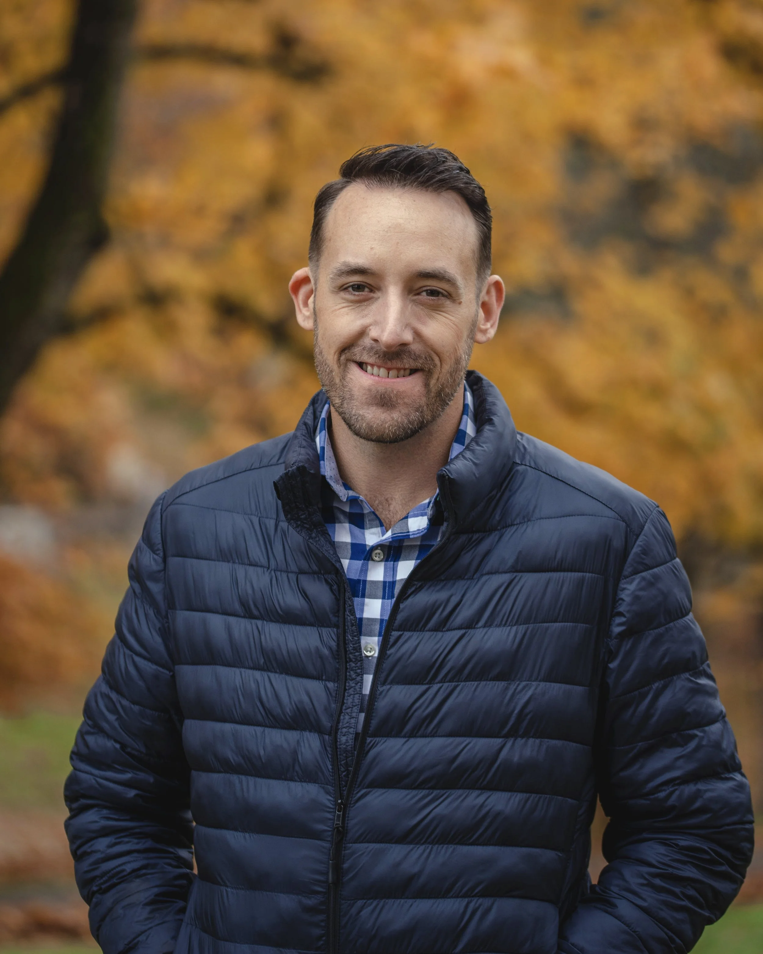 A smiling man outdoors in autumn, wearing a navy blue puffer jacket and a blue plaid shirt, with fall-colored leaves in the background.