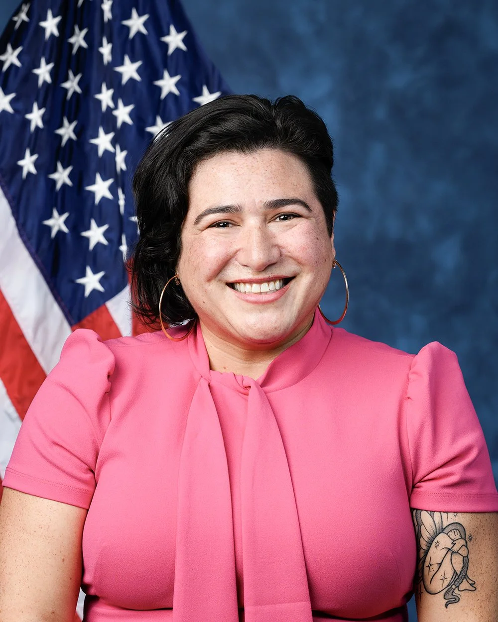 Smiling woman with dark hair, wearing a pink top with puffed sleeves and large hoop earrings, standing in front of an American flag.