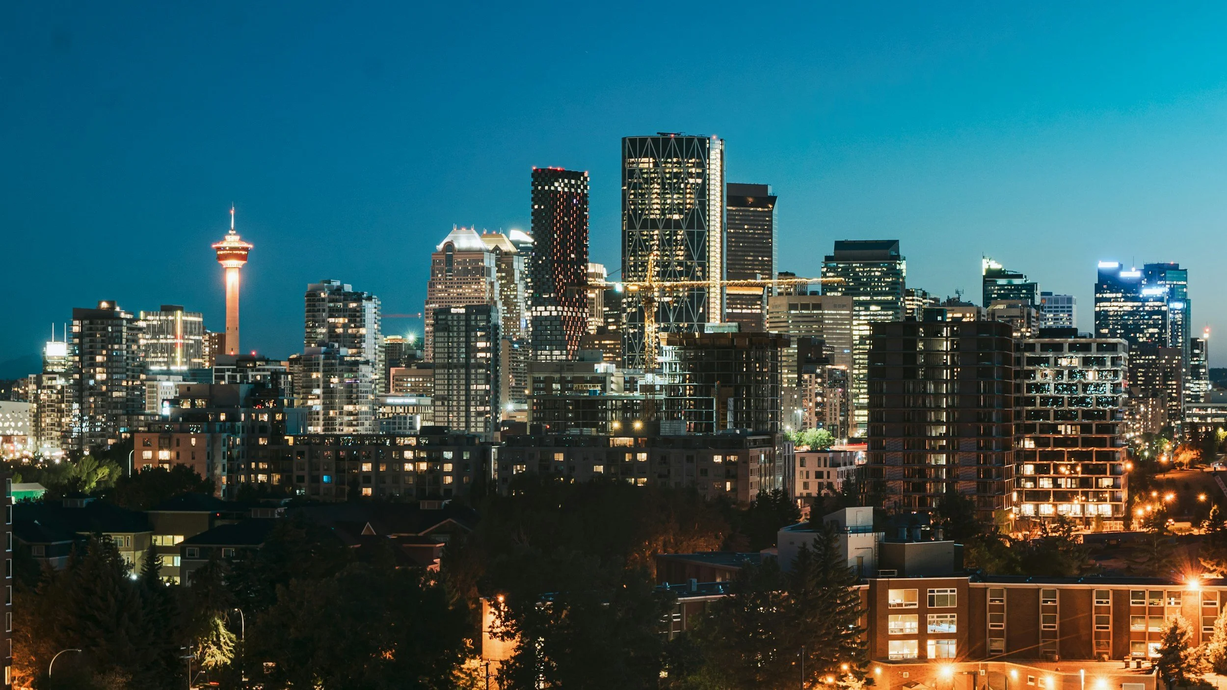 Nighttime city skyline with tall illuminated buildings and a tower with a sphere on top, under a clear dark blue sky.