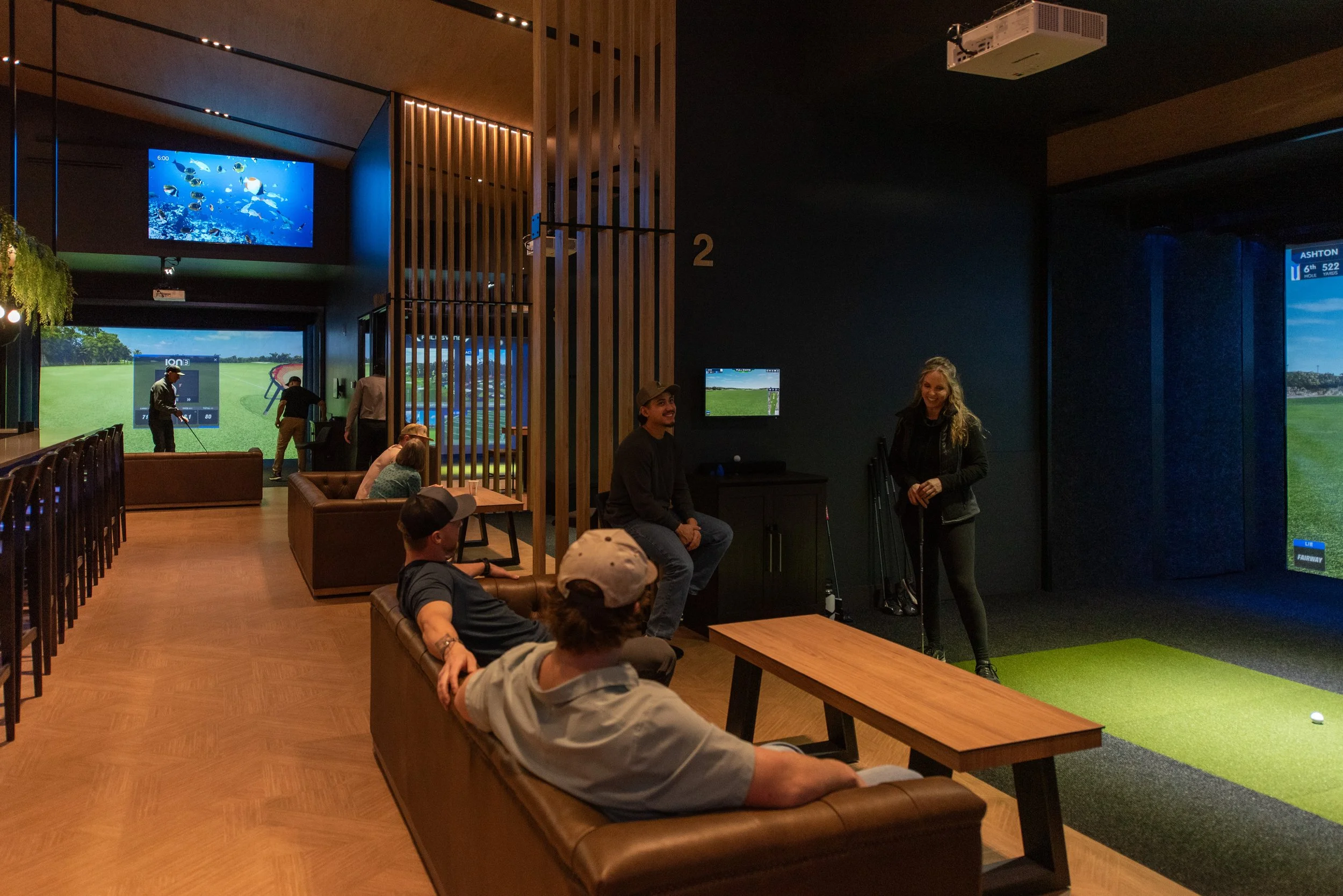 Guests enjoying indoor golf simulators at Arctic Fairways in Palmer, Alaska, with friends relaxing on couches while others play in private bays under warm modern lighting.
