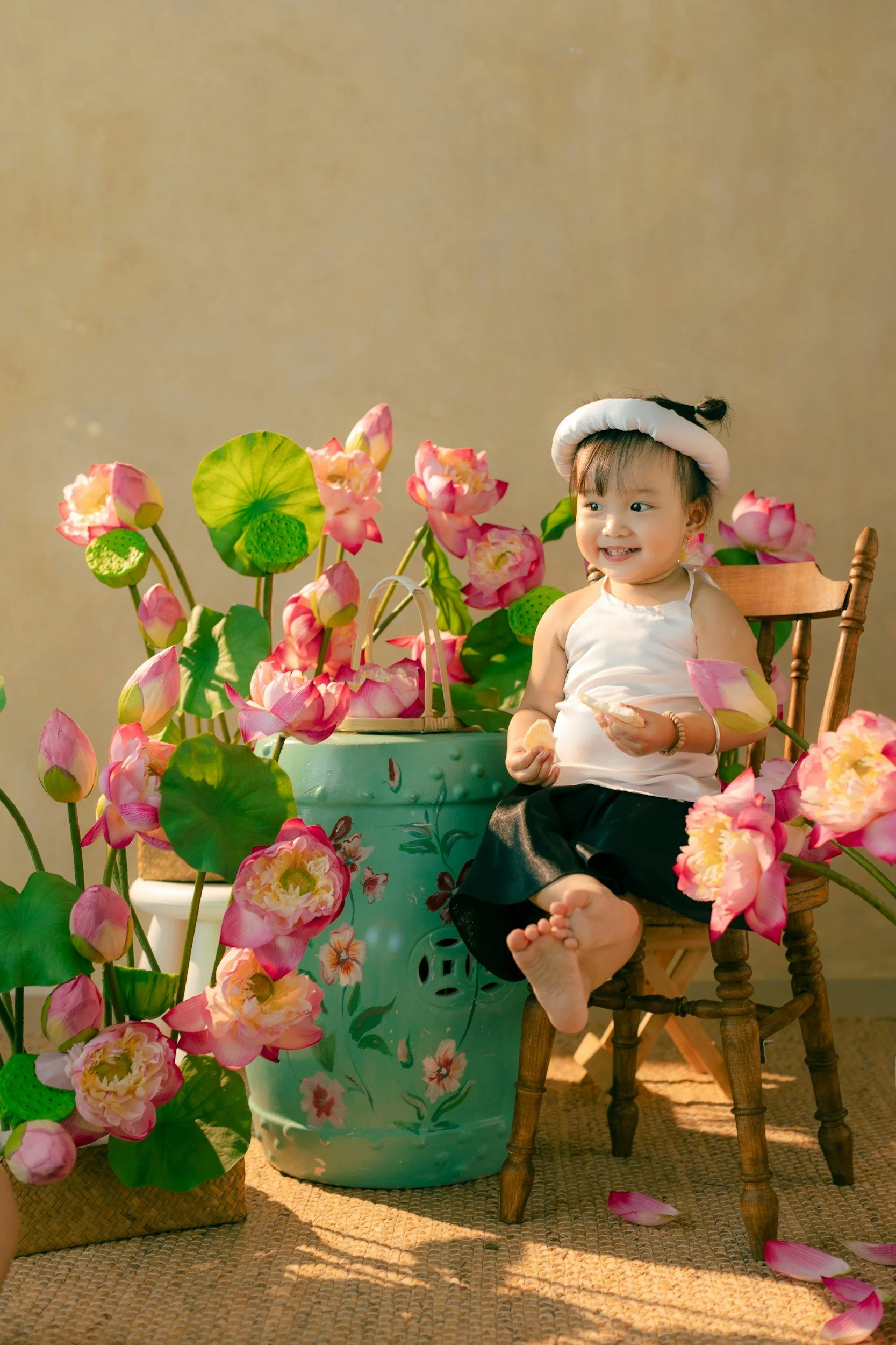 Smiling young girl sitting on a wooden chair, surrounded by pink lotus flowers and green foliage, holding a snack in each hand.