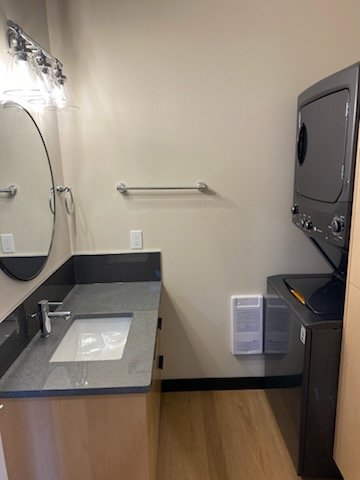 Bathroom with beige wall, gray countertop vanity with a sink, mirror, towel bar, and a stacked washer and dryer in corner.