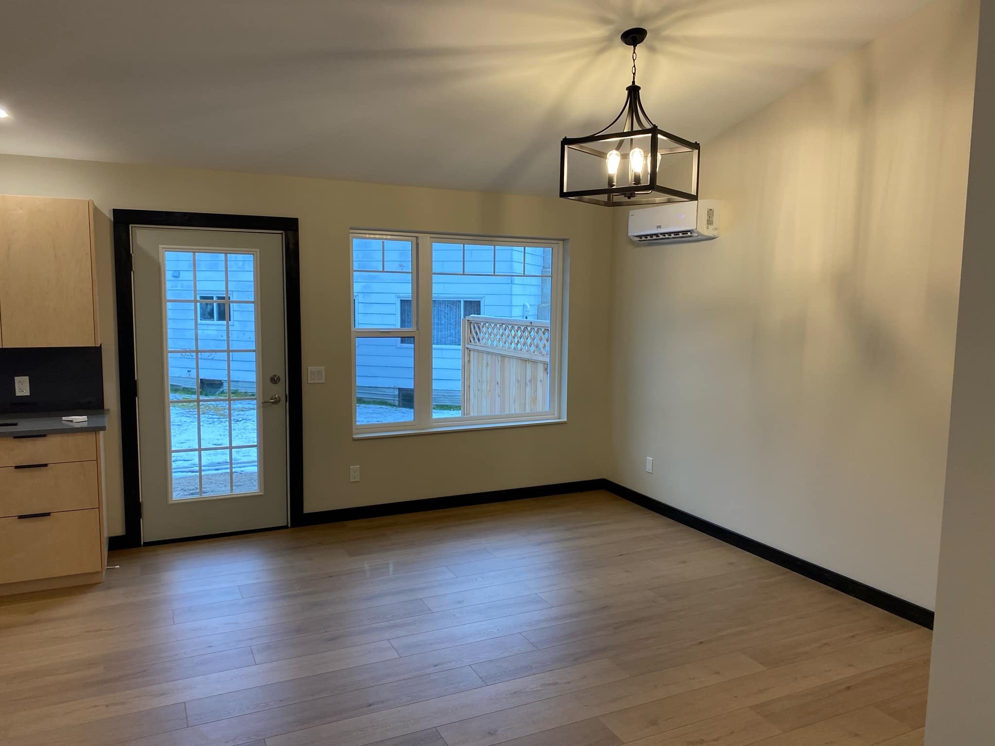 Empty living room with light wood flooring, cream walls, a black-framed door with glass panes, a window, a black ceiling light fixture, and a wall-mounted air conditioning unit.