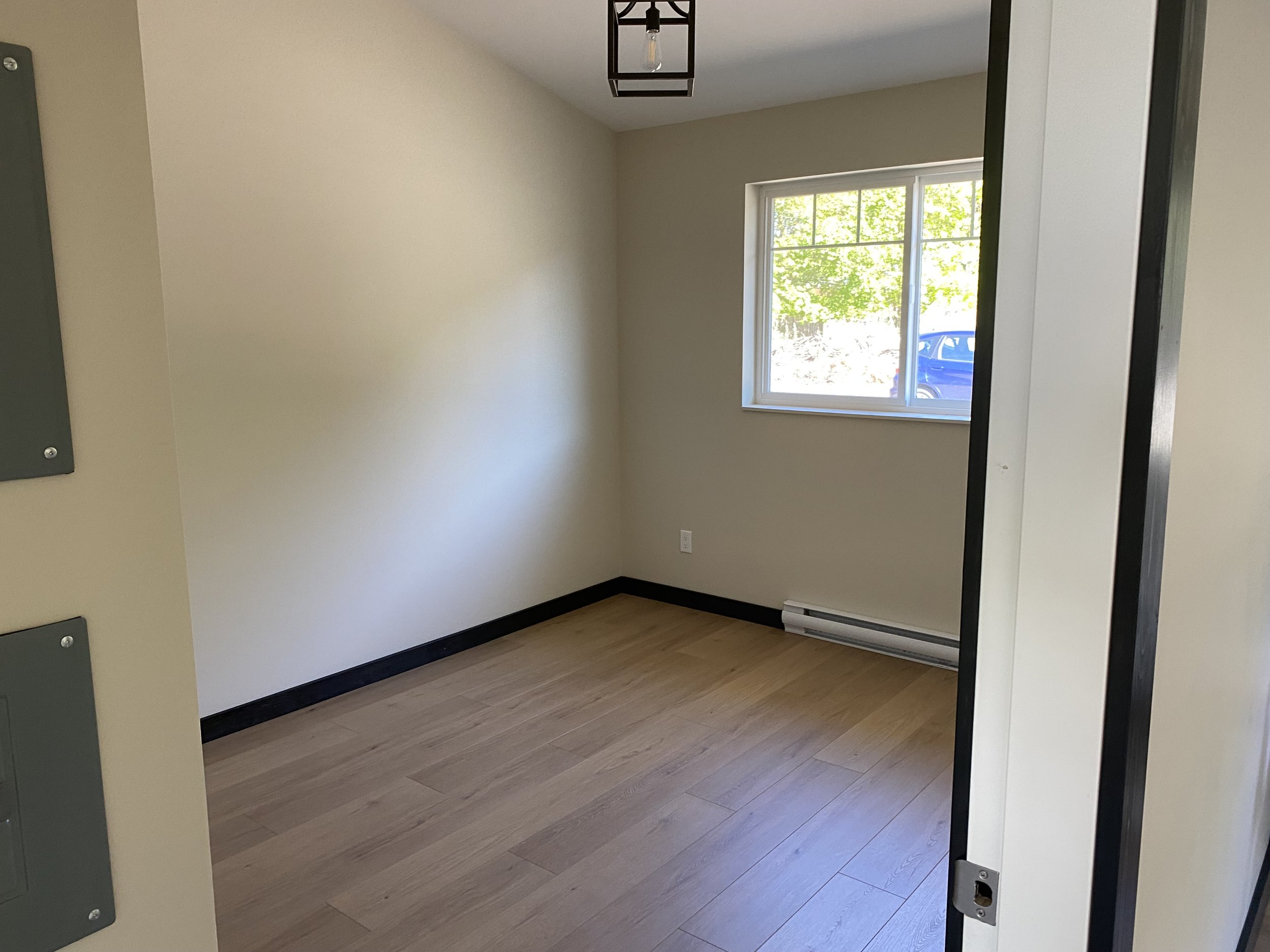 Empty room with beige walls, a window with green trees outside, light wooden floor, and a black baseboard heater.
