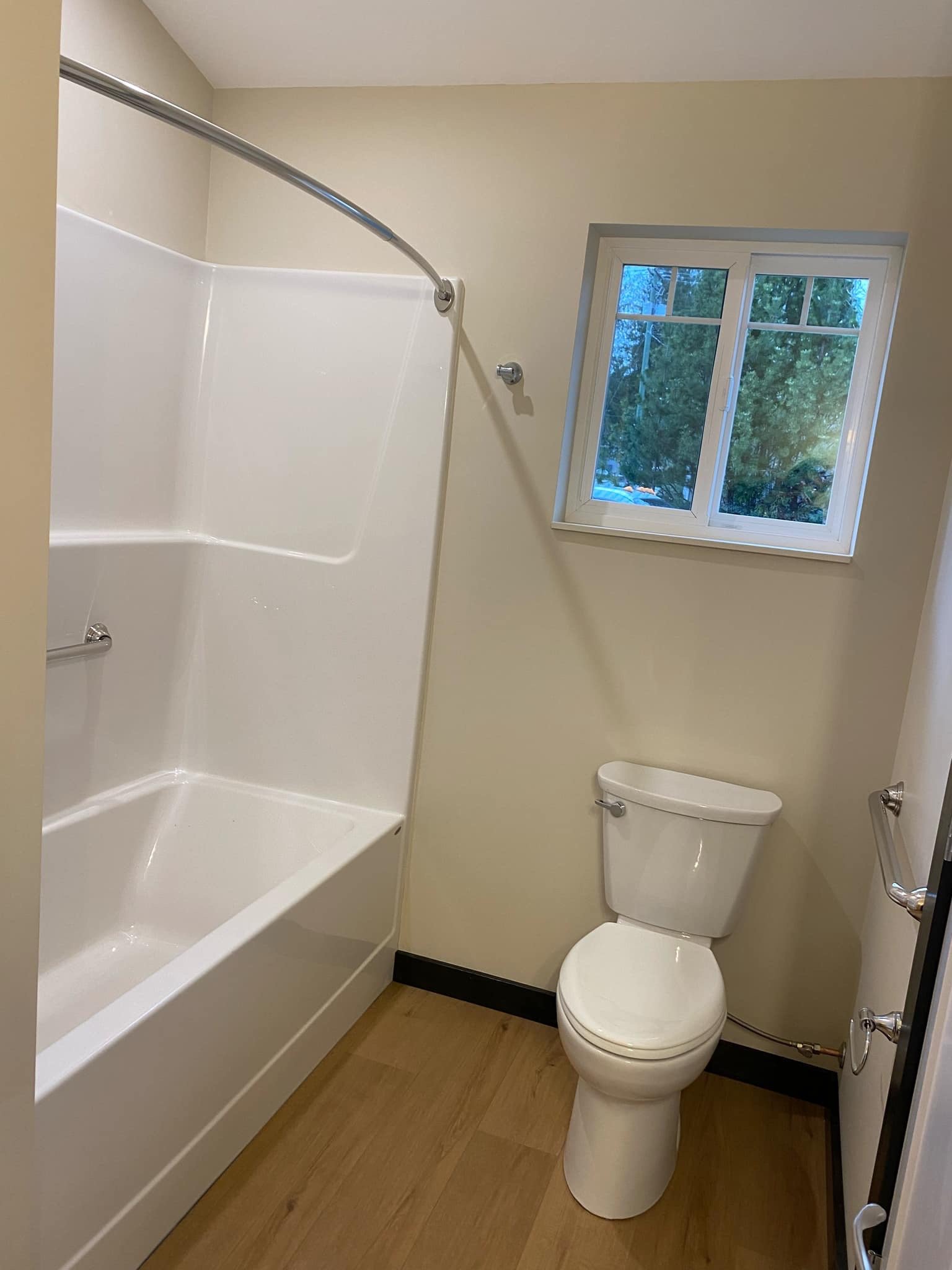A small bathroom with a bathtub and shower, a toilet, a window showing trees outside, and wood-style flooring.