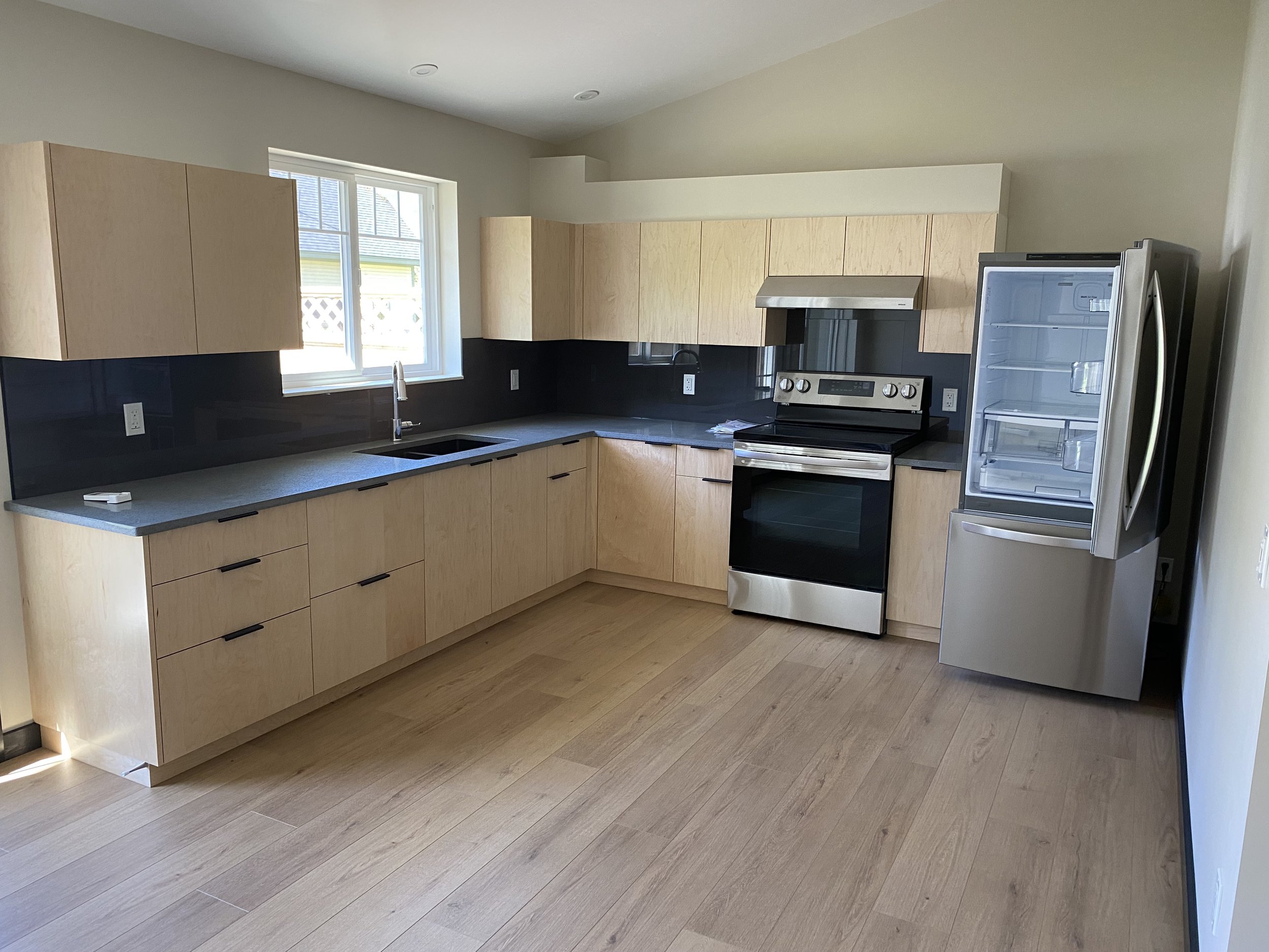 Empty modern kitchen with light wood cabinets, gray countertops, a black backsplash, stainless steel appliances, and a window above the sink.