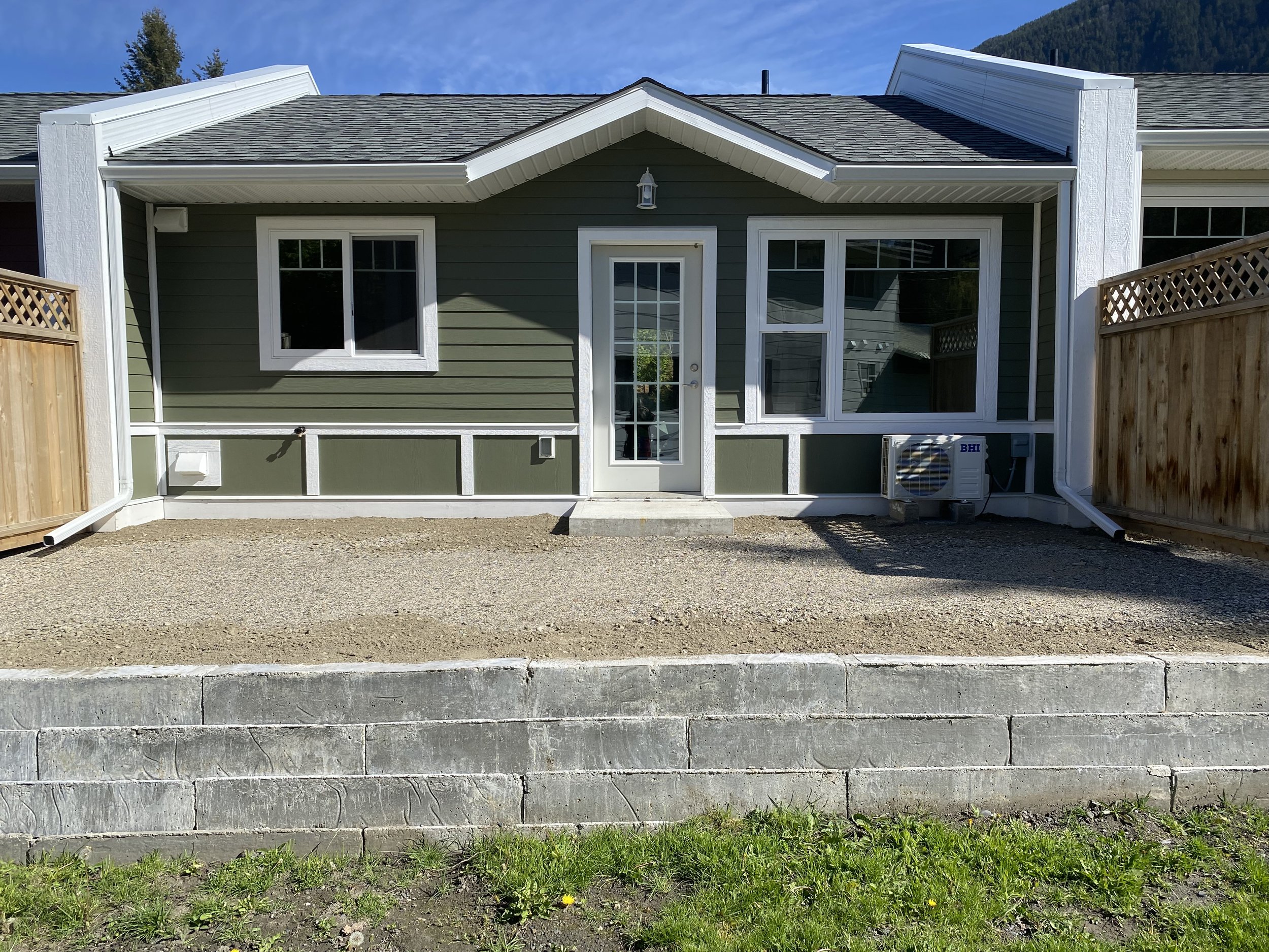 Back of a green house with white trim, two front windows, a glass door, an air conditioning unit, and a wooden fence on each side, with a gravel and dirt yard and a concrete retaining wall in front.