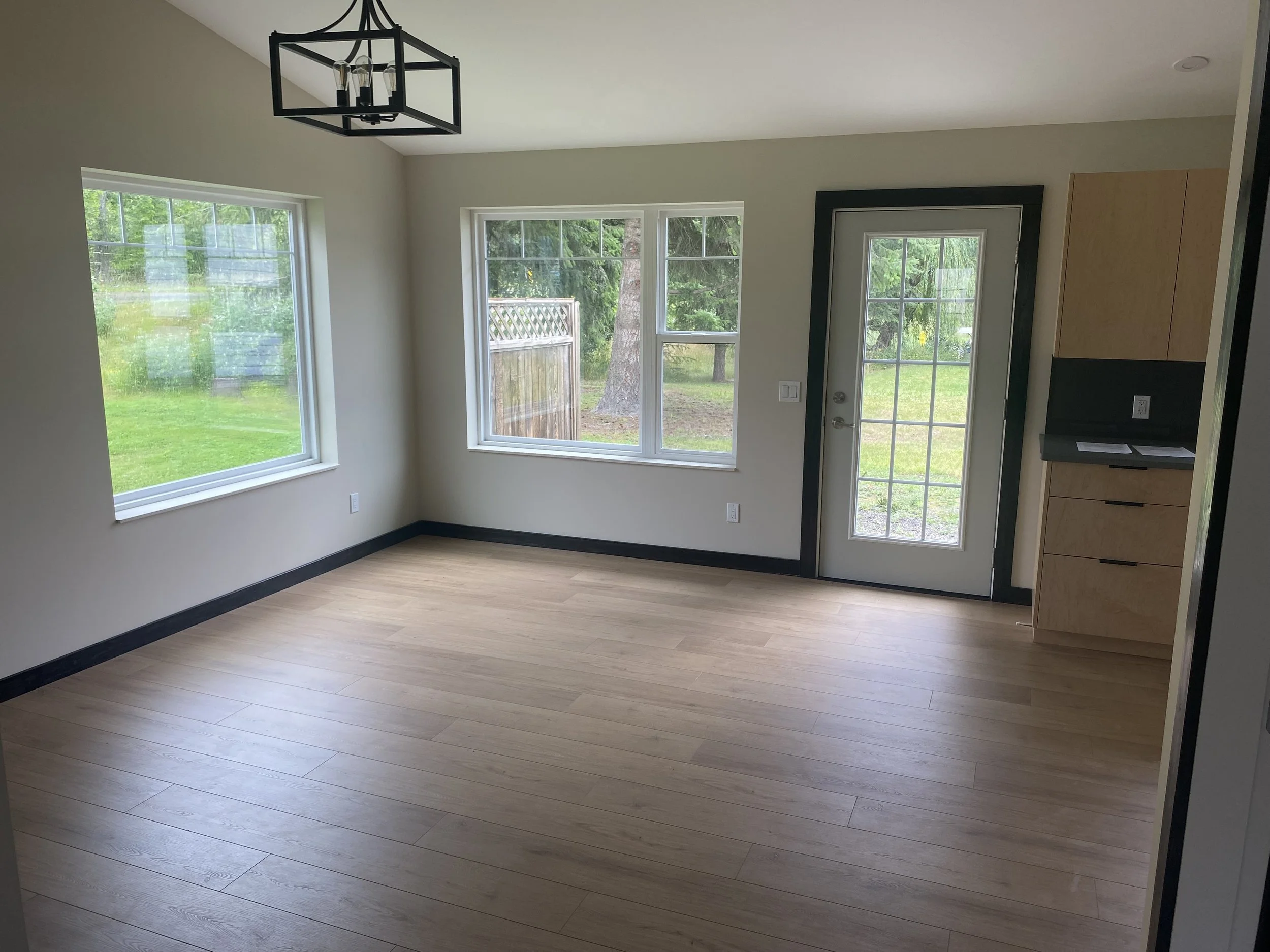 Empty room with light-colored wooden flooring, white walls, large windows, a glass door leading outside, and a black-framed chandelier