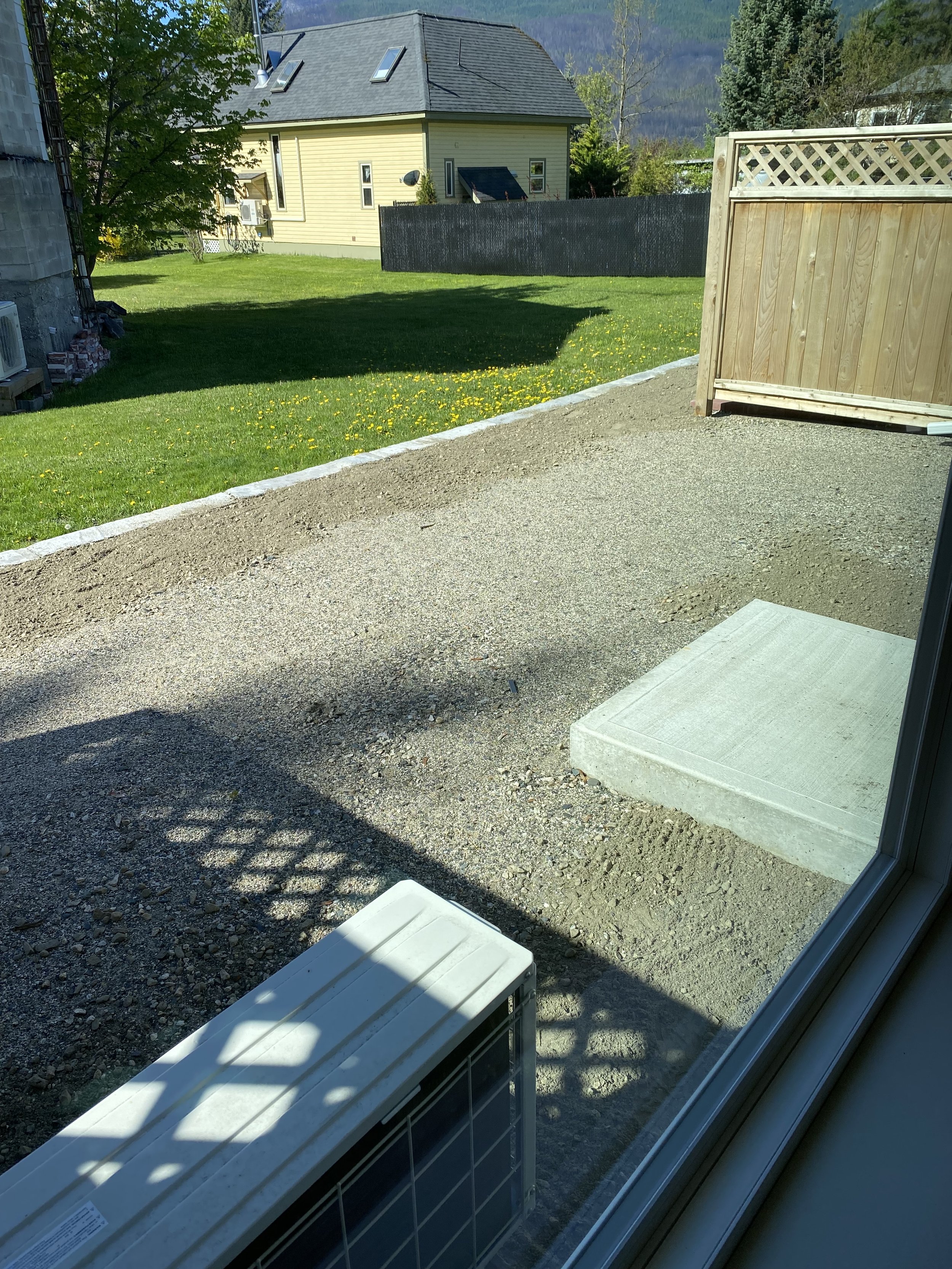 View through a window of a backyard with a gravel patio, a new concrete step, and a wooden fence. Green grass with yellow dandelions, a yellow house with a dark roof, trees, and mountains in the distance.