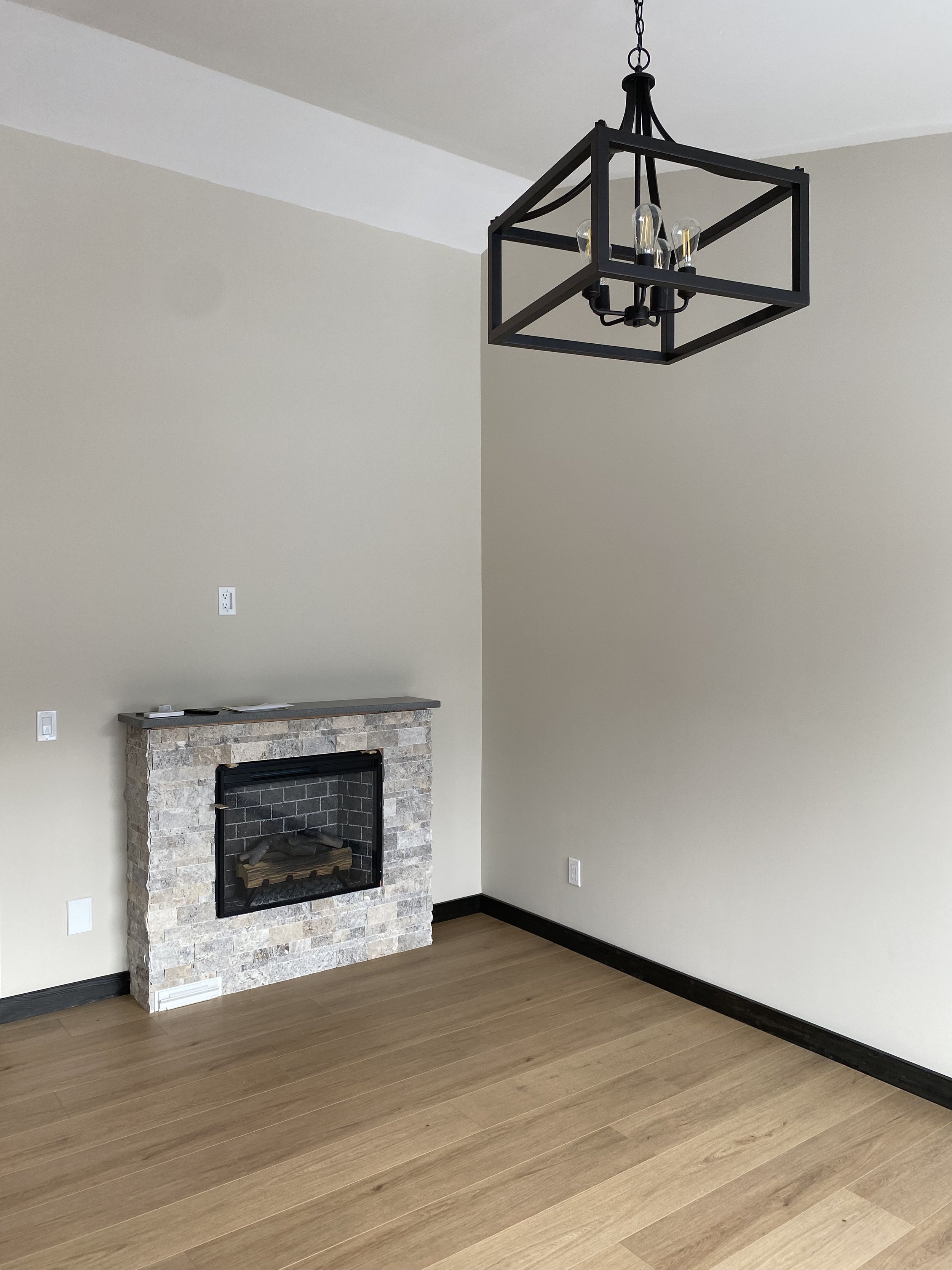 Empty living room with a stone fireplace, wooden floor, beige walls, black baseboards, a black metal ceiling chandelier, and electrical outlets.
