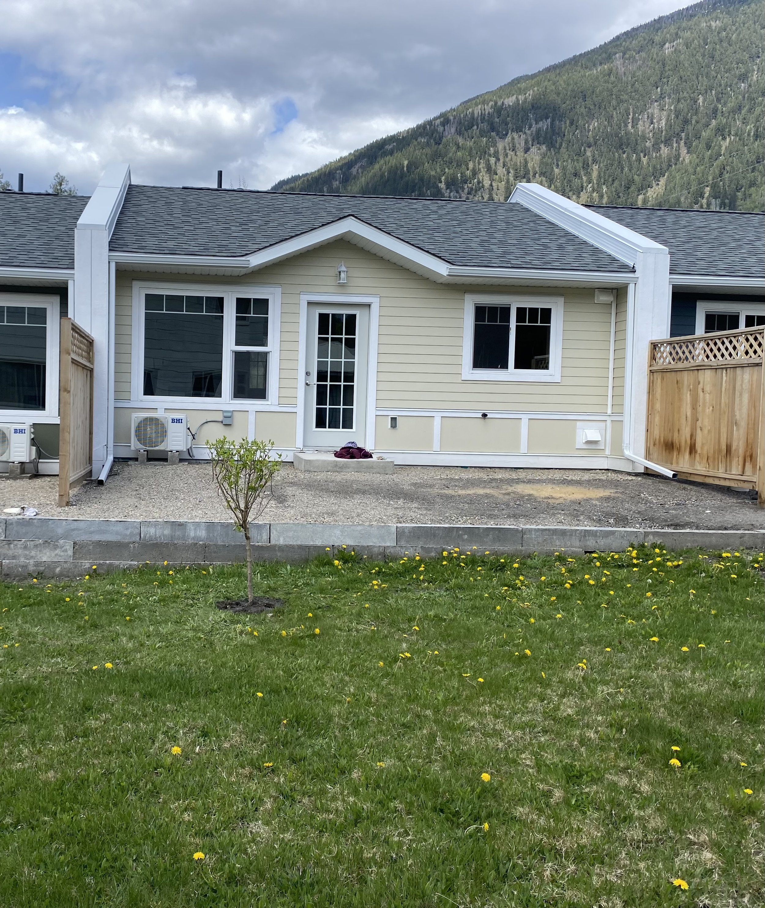Backyard view of Creekside Villas in New Denver, BC with a small tree in the grass, a patio area with gravel, and a mountain in the background.