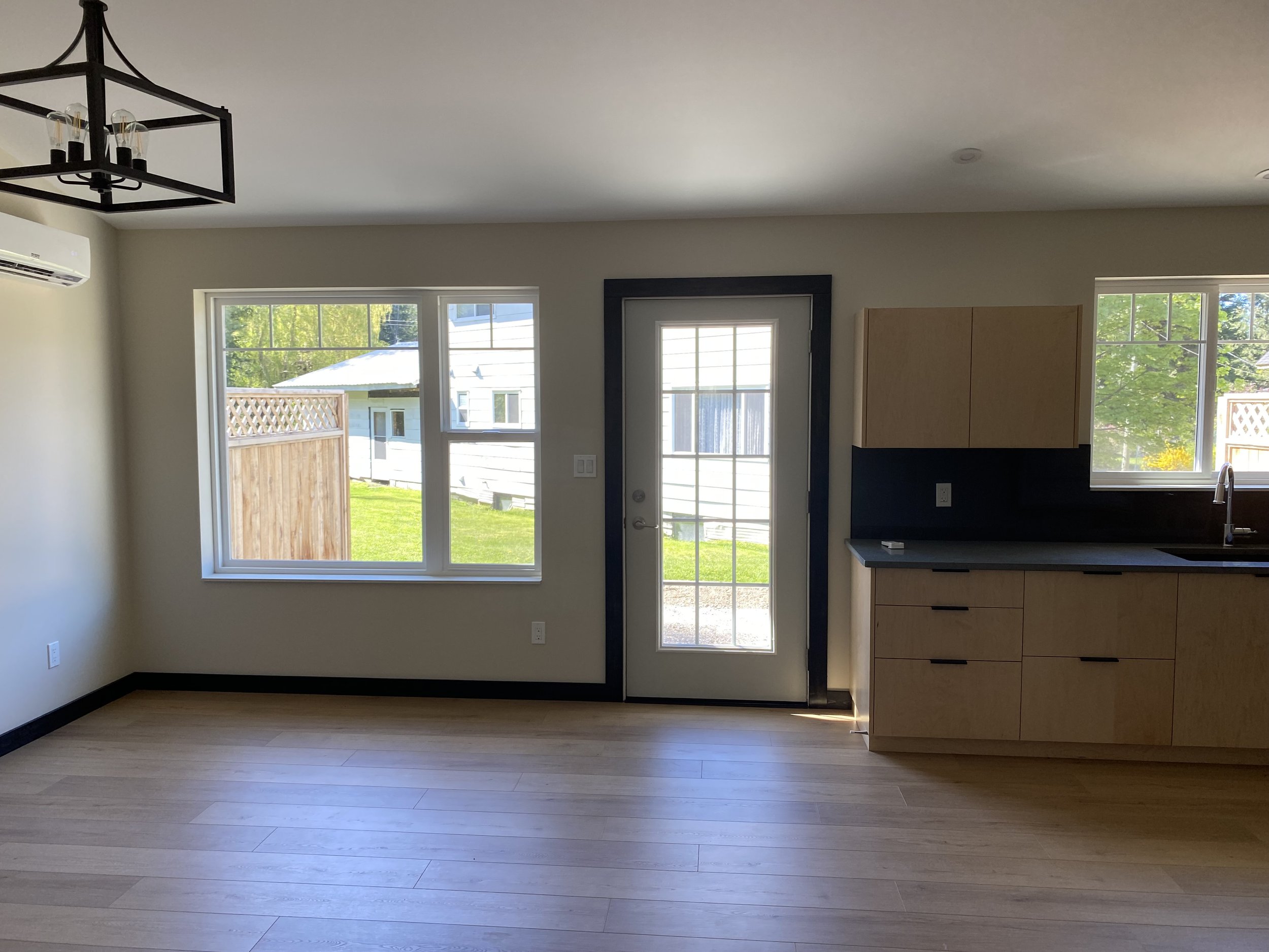 Empty kitchen and dining area with large windows, a door with glass panes, light wood flooring, and new cabinets in a house.
