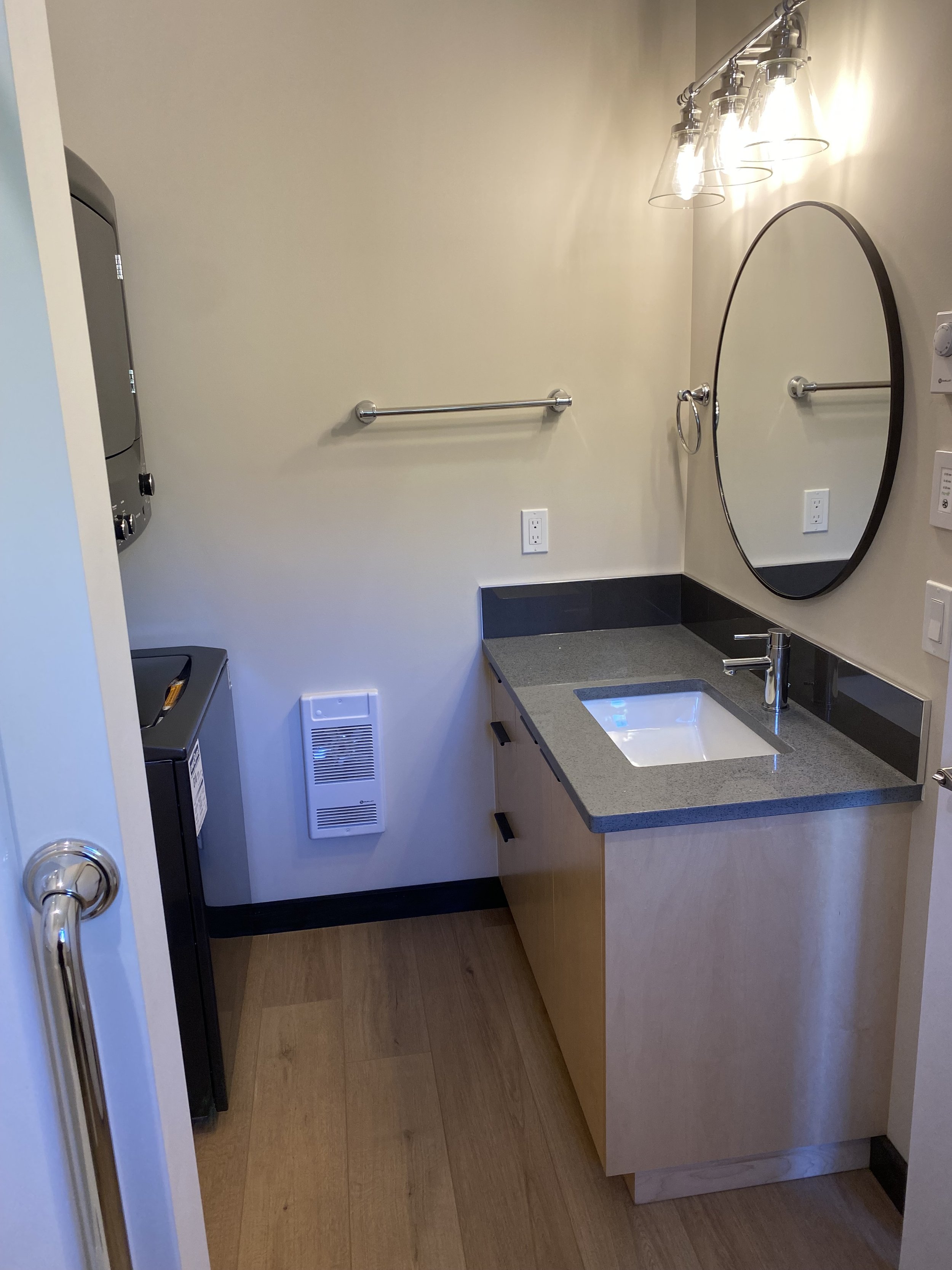 Small bathroom with beige walls, a wooden cabinet with a grey countertop and built-in sink, a round mirror, wall-mounted light fixtures, a towel bar, laundry appliances on the left, and a wall heater.
