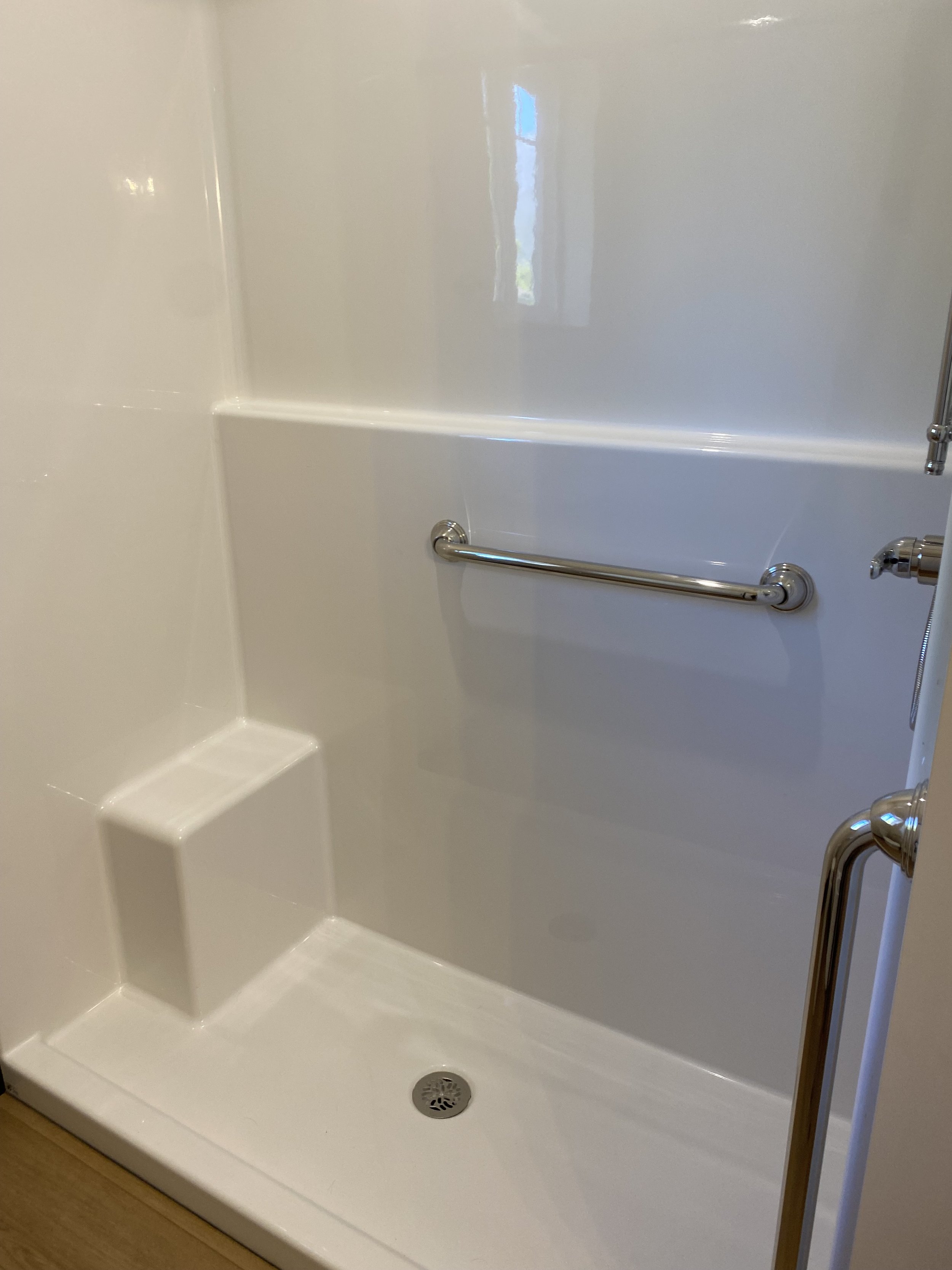 Empty white shower with a horizontal grab bar and a built-in corner seat, with a drain in the floor.