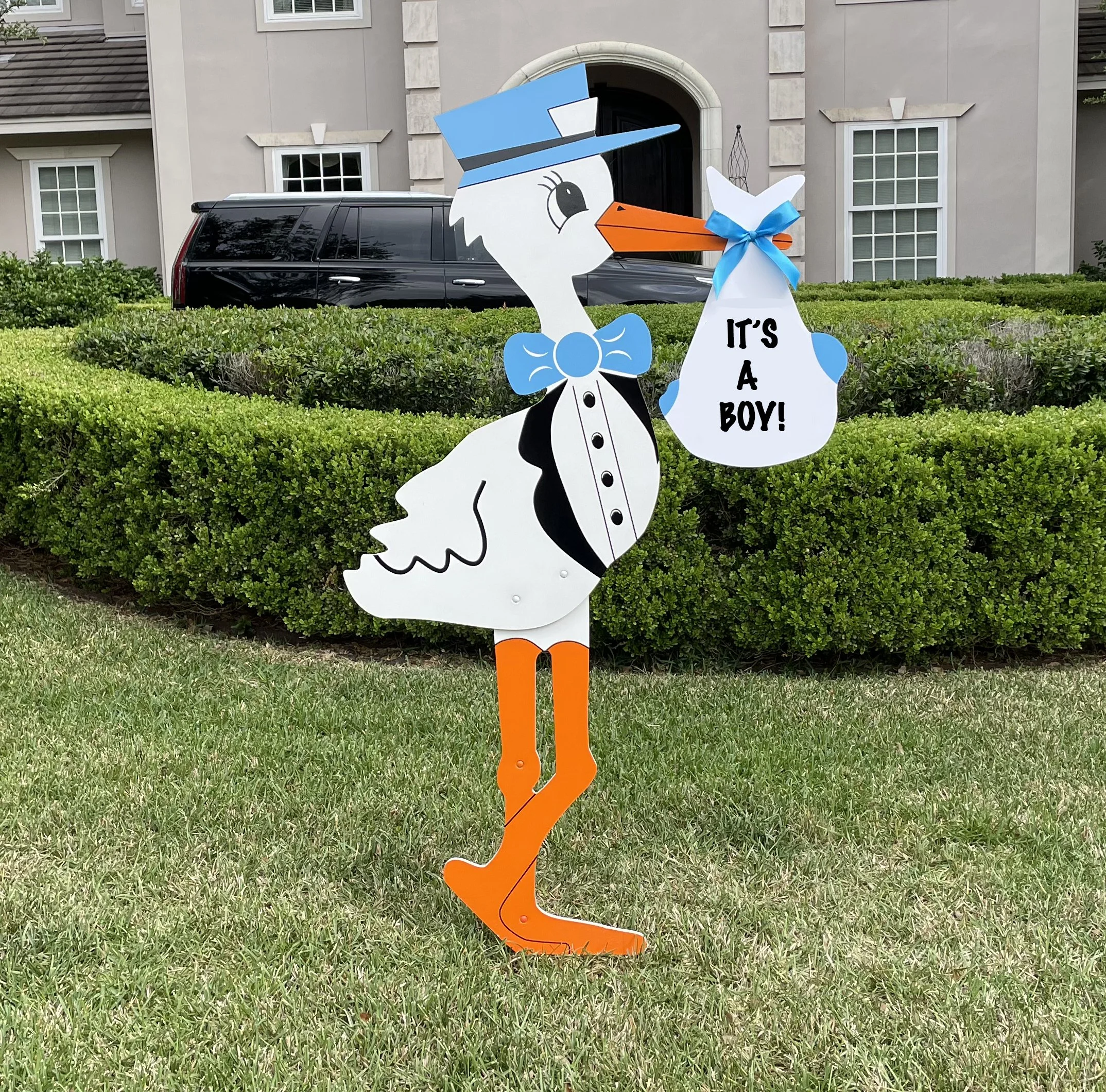 Yard decoration of a cartoon stork holding a bag that says 'It's a Boy!', standing on grass in front of a hedge and house.